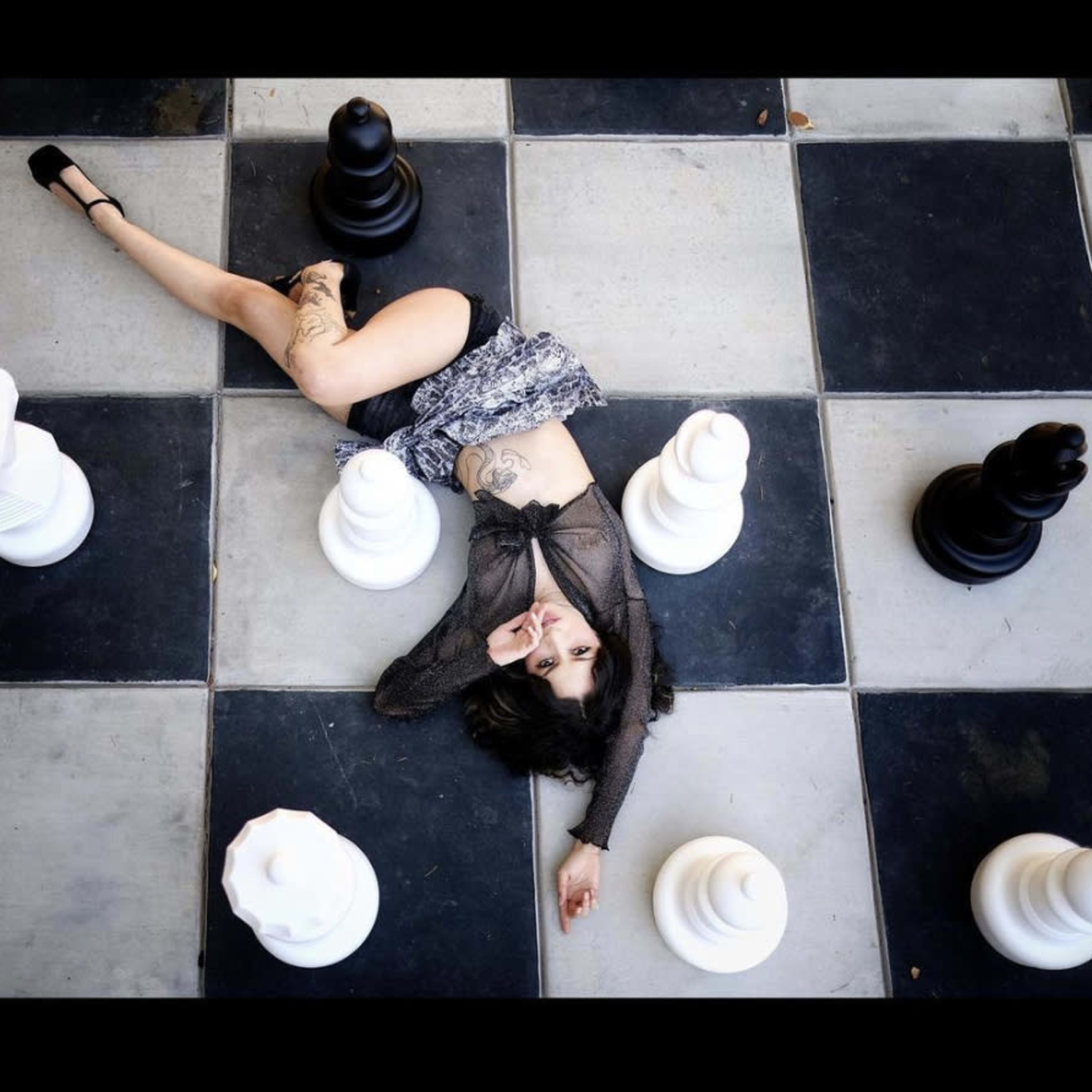 A person lies on a large checkered chessboard, surrounded by oversized black and white chess pieces.