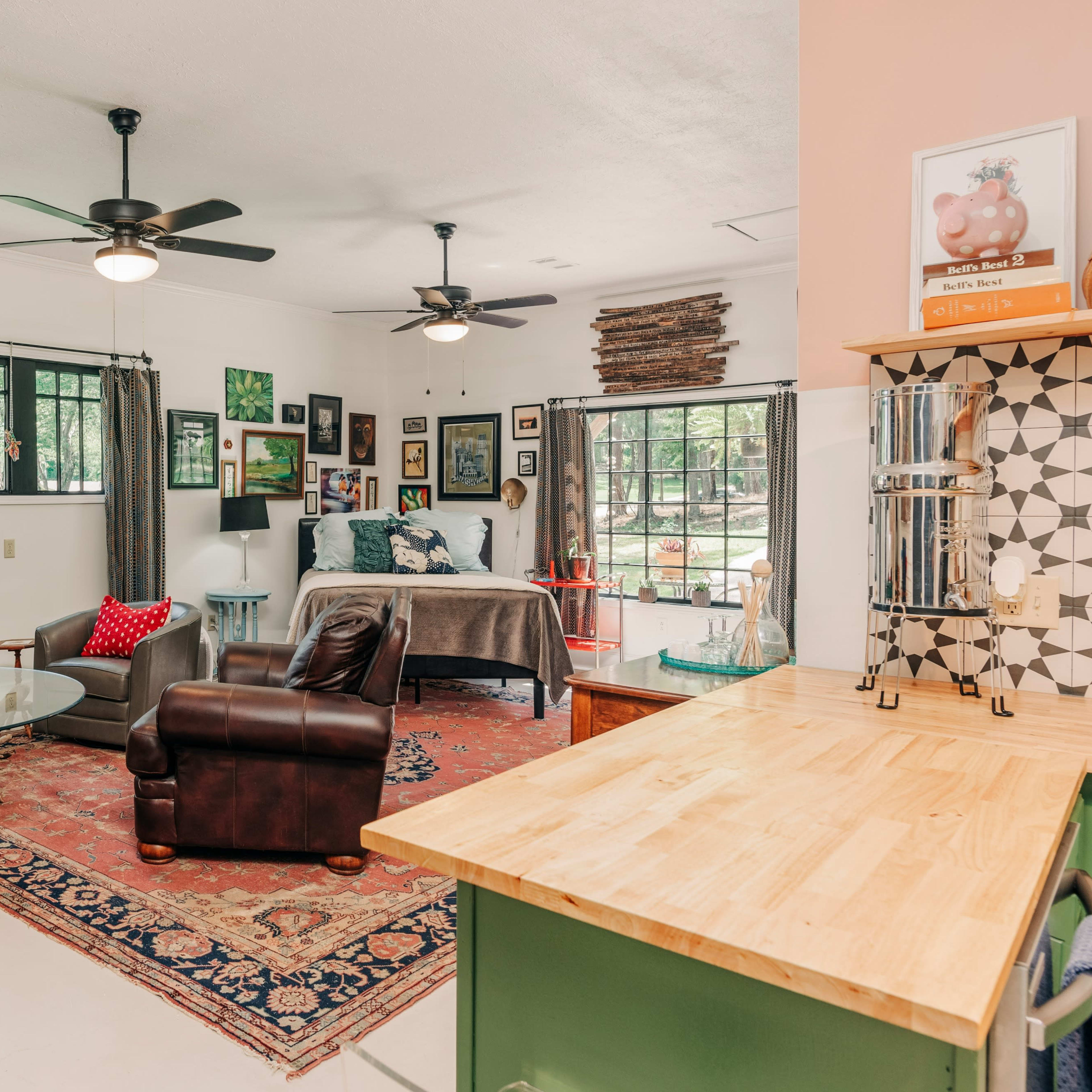 A spacious room features a living area with a brown leather chair, a dining table, and large windows that overlook an outdoor view, with various artworks displayed on the walls.