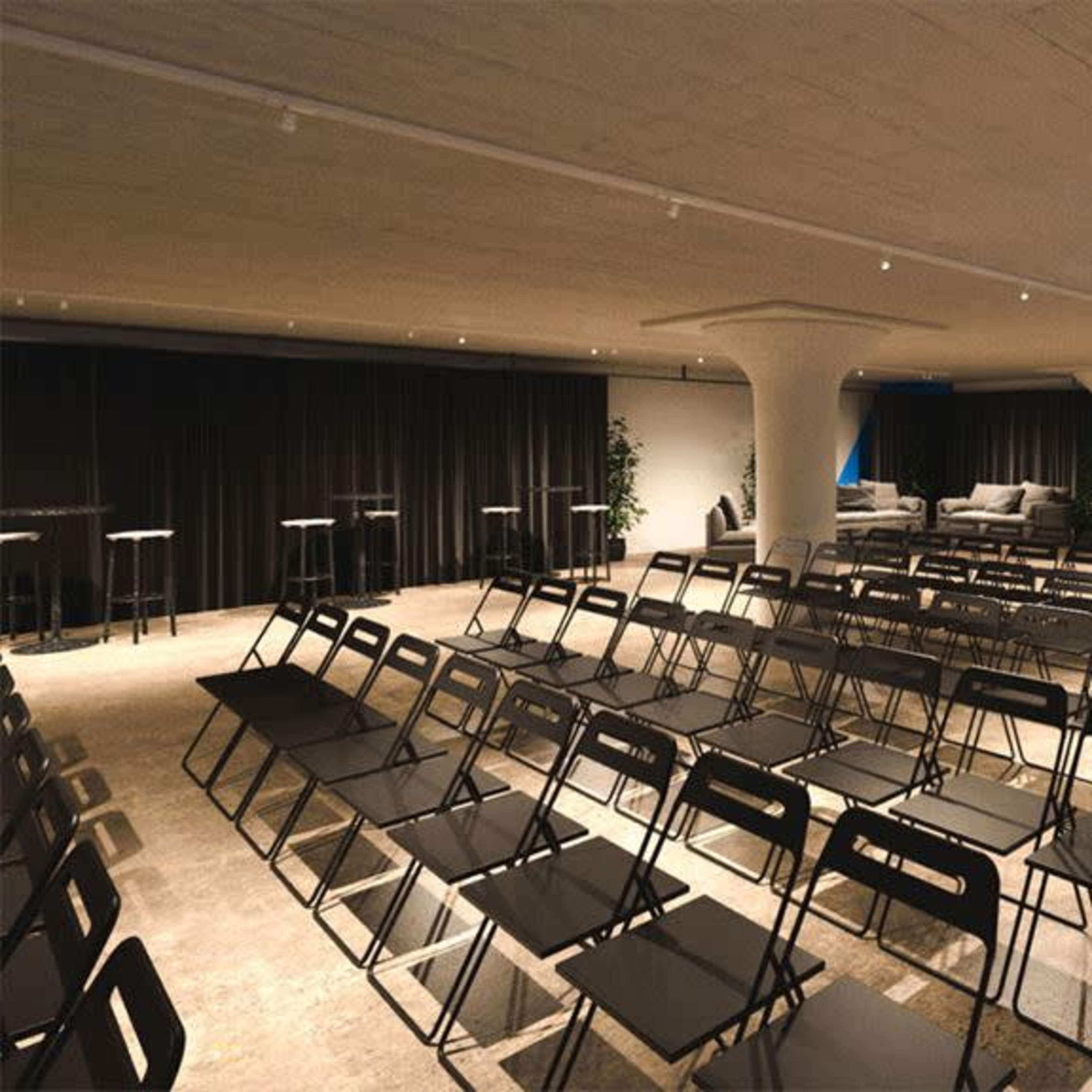 A modern event space with rows of black chairs facing a seating area, illuminated by natural light and featuring blue architectural highlights.