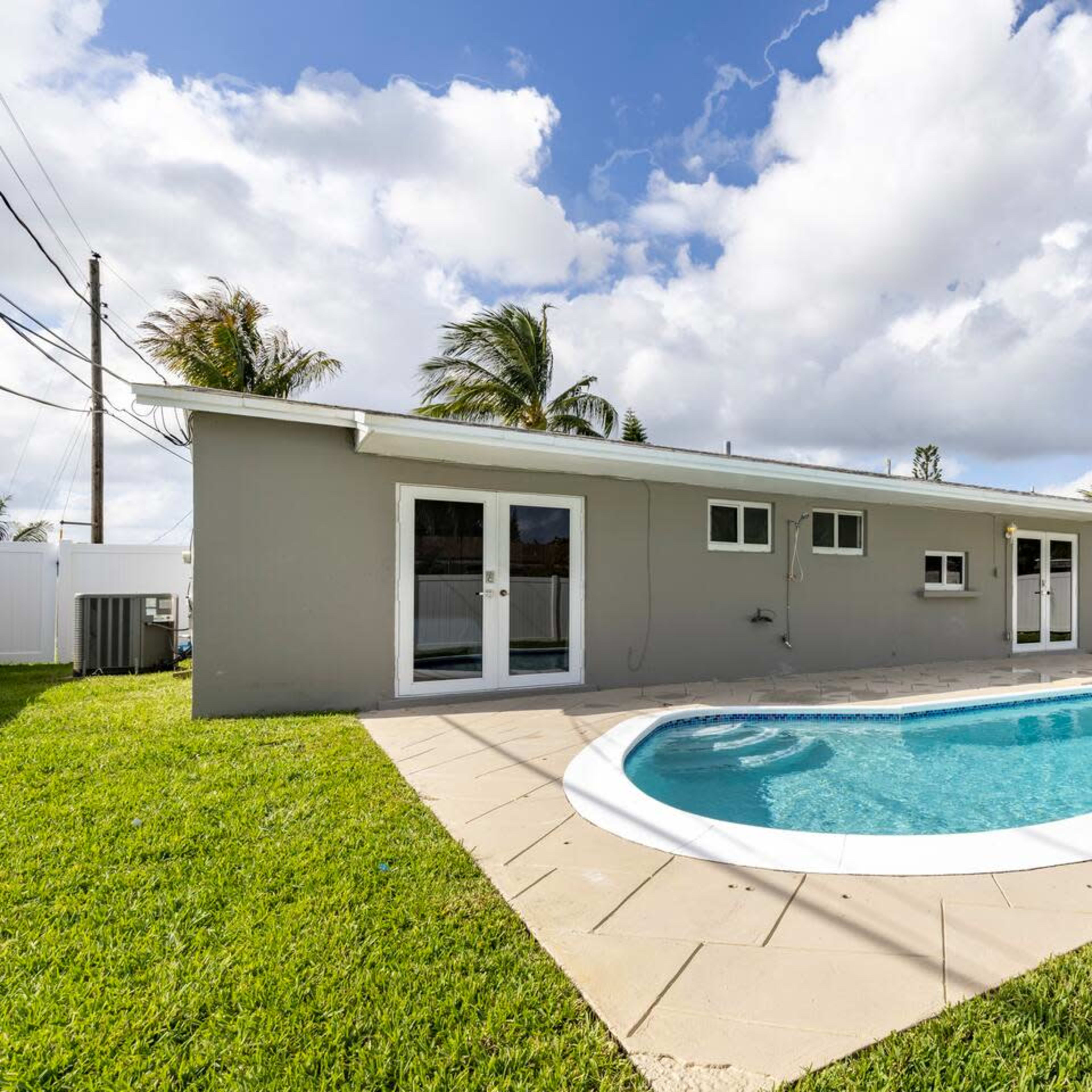 A rectangular swimming pool is set in a backyard next to a single-story house with sliding glass doors and surrounded by well-maintained grass and palm trees.