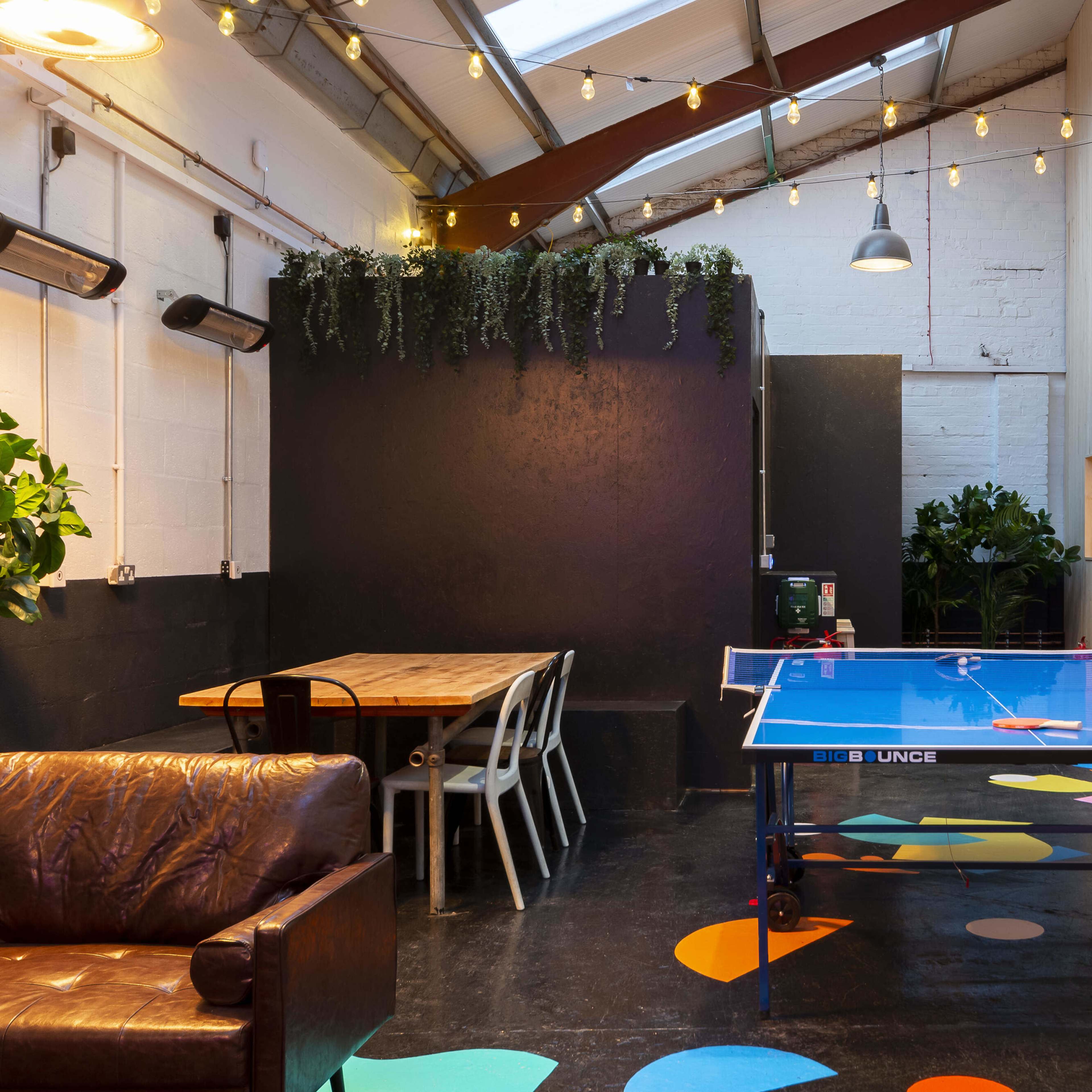 A brightly lit indoor space featuring a blue ping pong table, wooden seating, and several numbered rooms along the wall, decorated with plants and colorful floor designs.