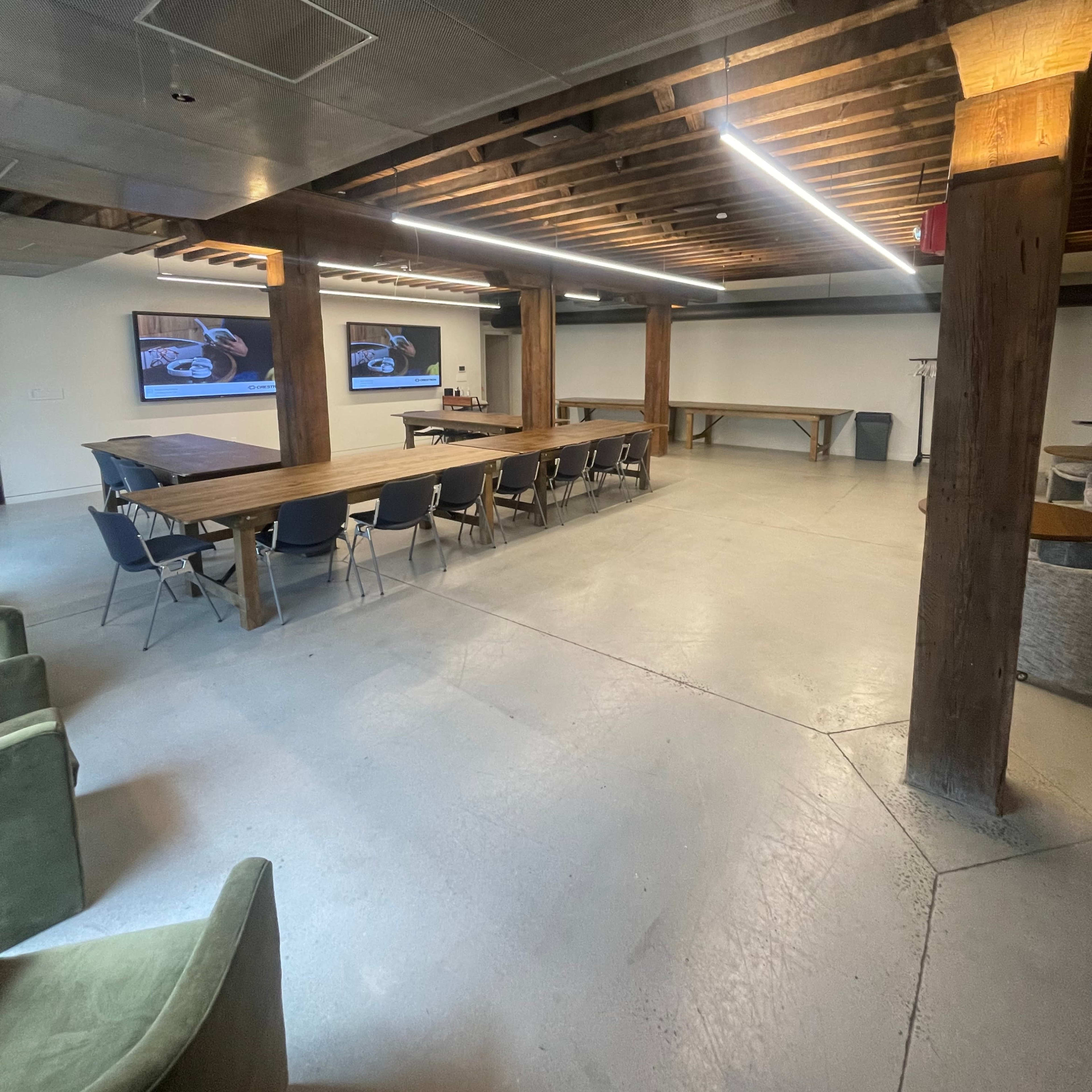 A spacious meeting room with wooden beams, a large table, rows of chairs, and two screens mounted on the walls.