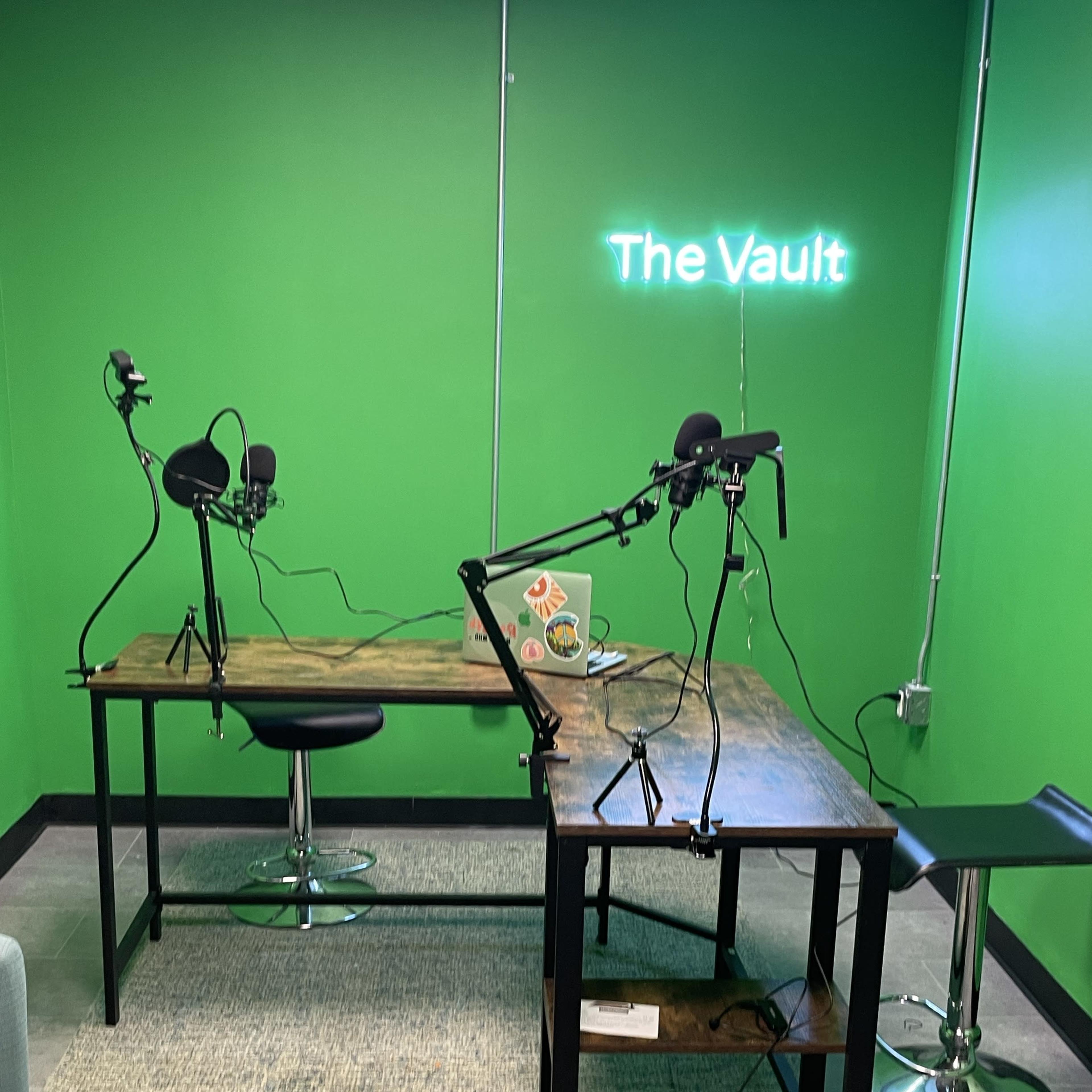 A podcast studio features a wooden table with two microphones on stands, green walls, and a neon sign reading "The Vault."