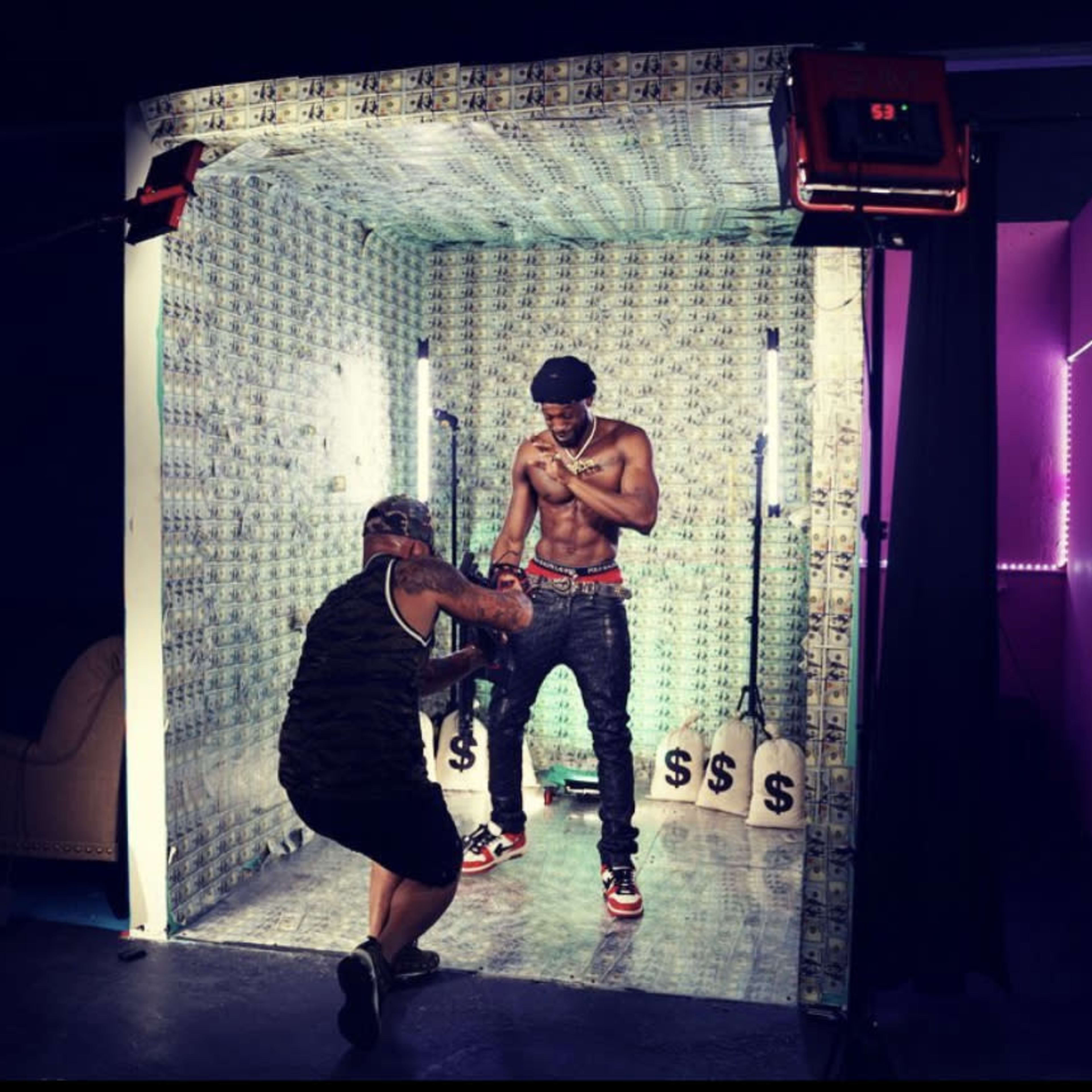 A shirtless man poses inside a room decorated with money-themed walls while another person adjusts the lighting.