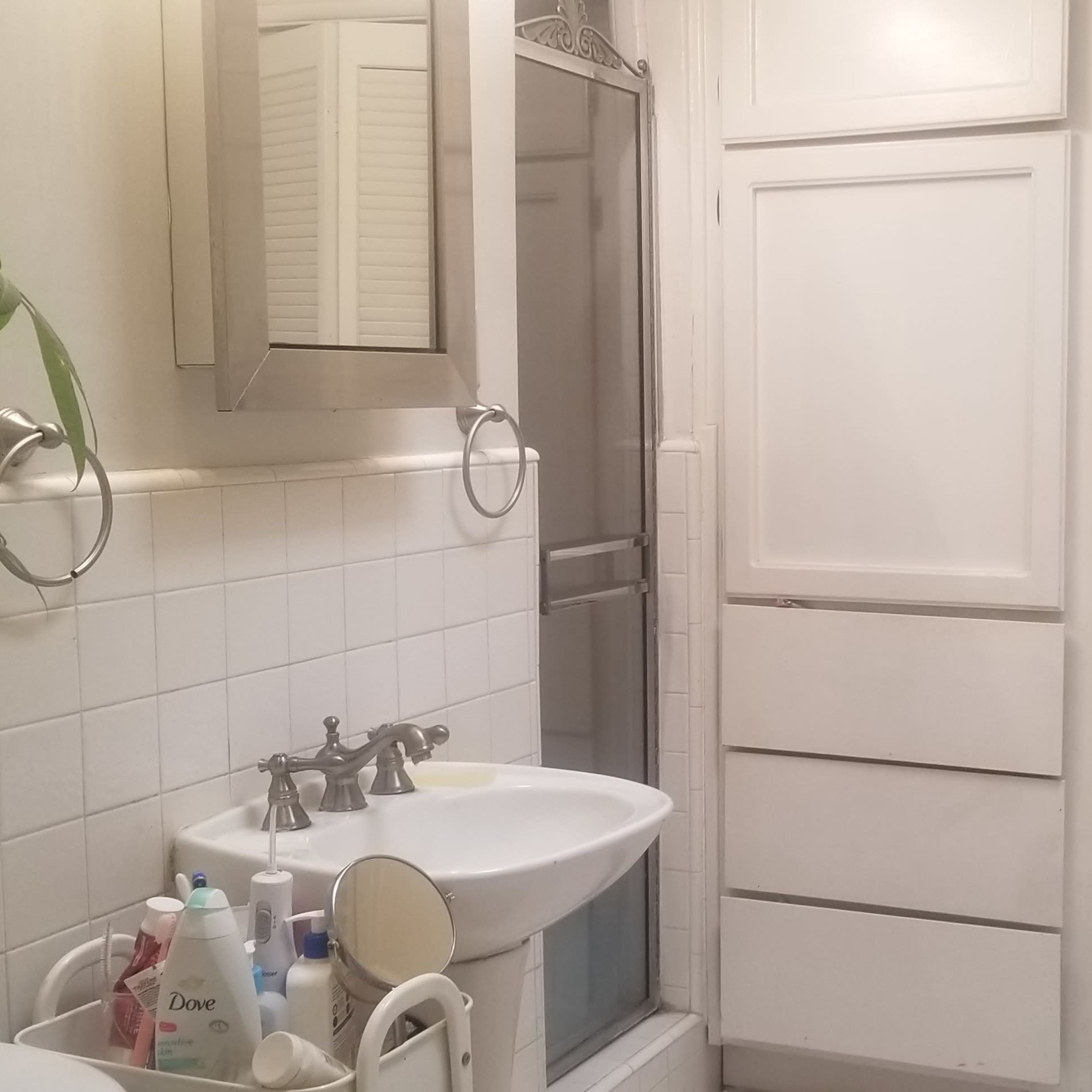 A small bathroom features a sink, a mirror, and white cabinetry, with a shower and a chair positioned nearby.
