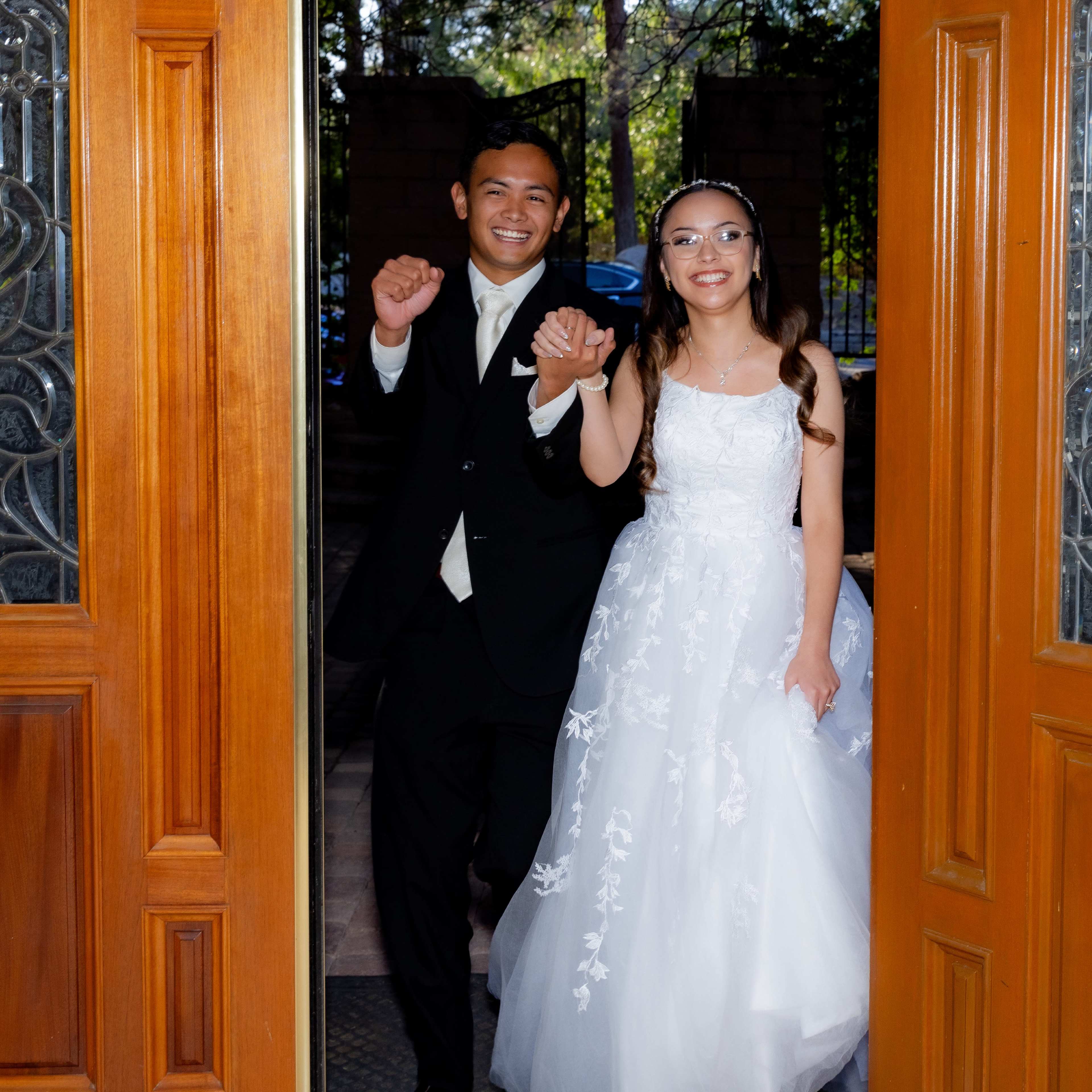 A young couple exits through a wooden door, both smiling and holding hands, with the woman wearing a white wedding dress.