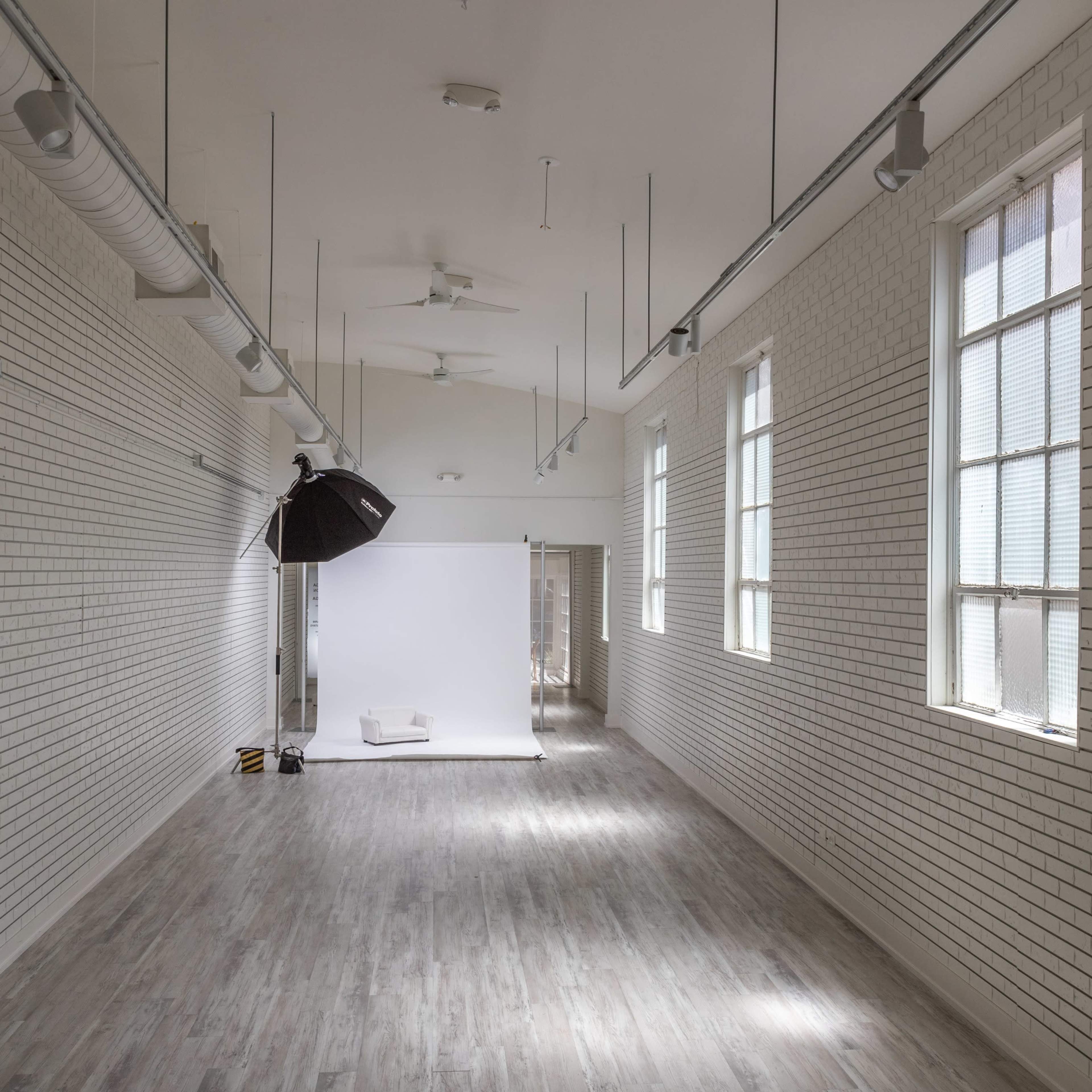 A long, narrow studio space with white brick walls, large windows, and a backdrop setup at one end.