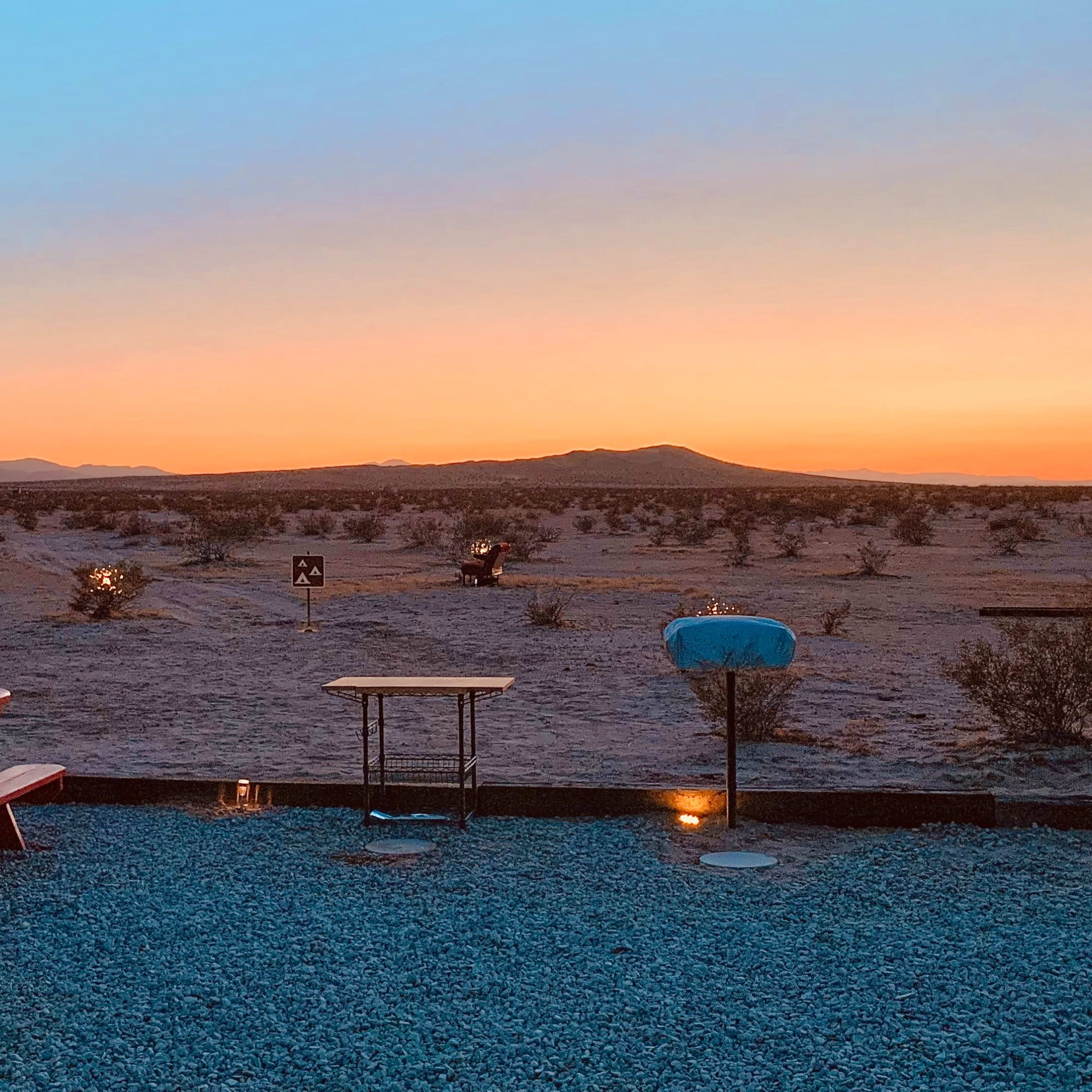 A picnic table sits on gravel in a desert landscape under a sunset sky with silhouetted mountains in the background.