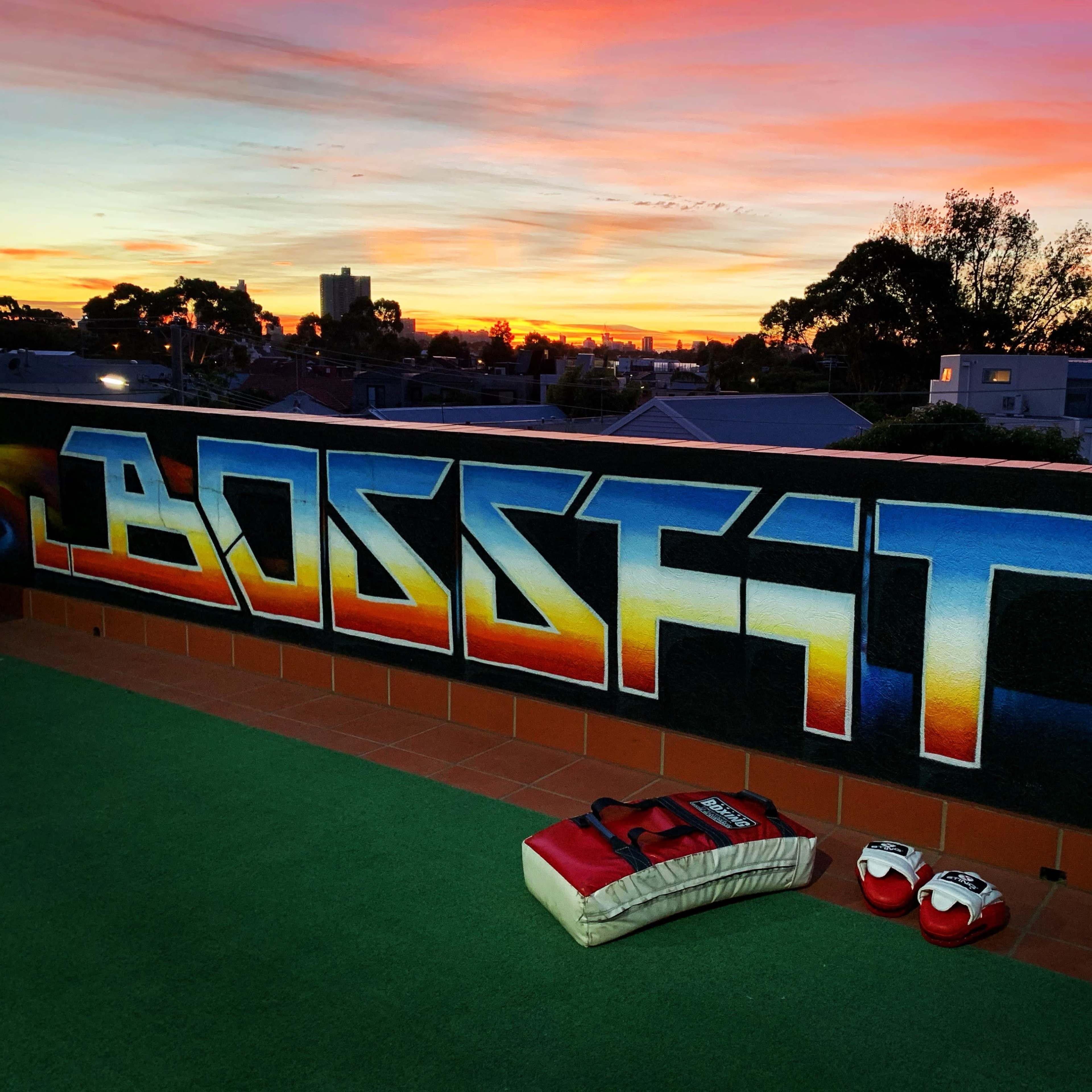 A colorful sunset over rooftops, with a mural reading "BOSSFIT" and boxing gloves on a green surface in the foreground.