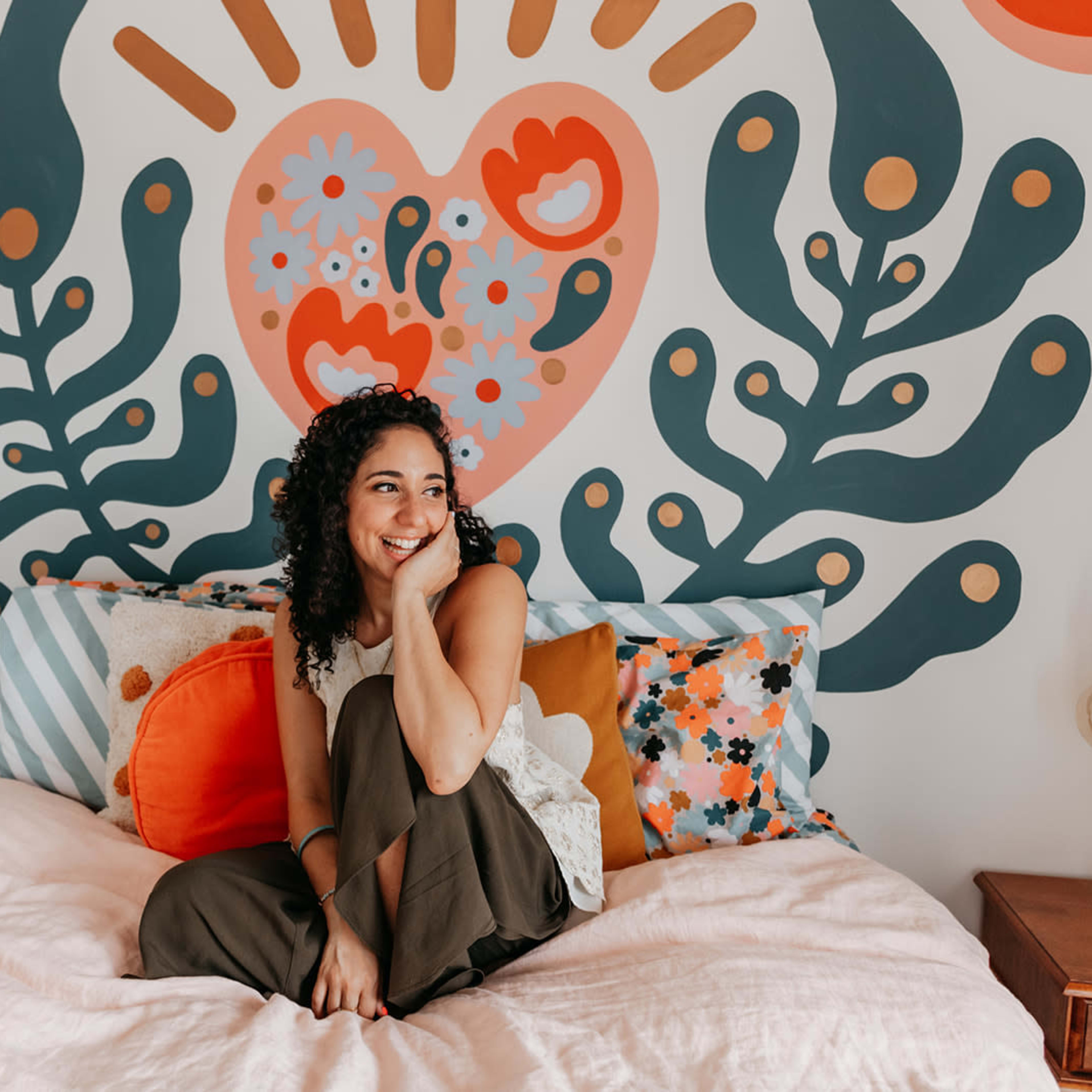 A woman with curly hair sits on a bed adorned with colorful pillows in front of a large, patterned mural on the wall.