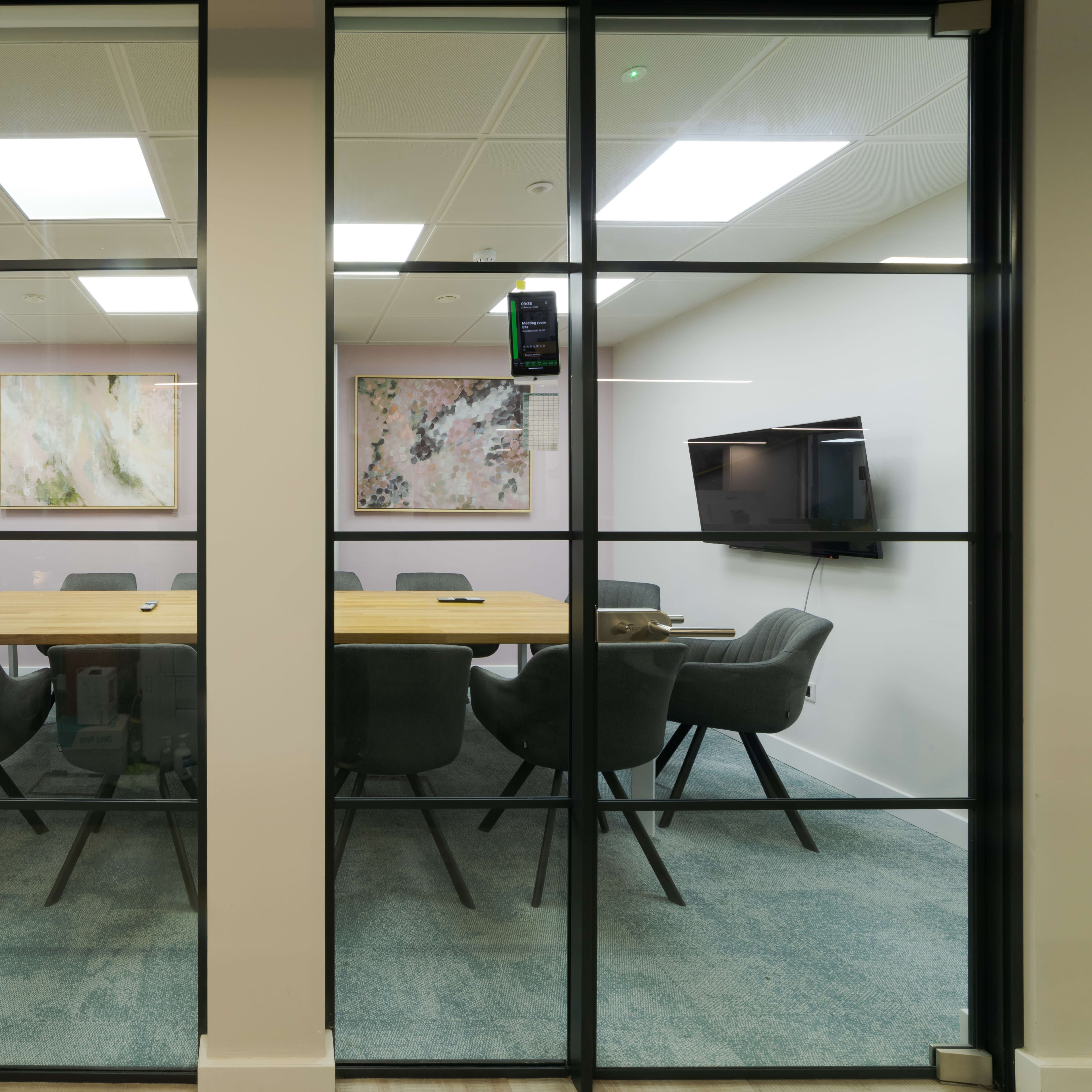 A modern conference room with a long wooden table surrounded by gray chairs is visible through glass walls, featuring two wall-mounted televisions and abstract artwork.