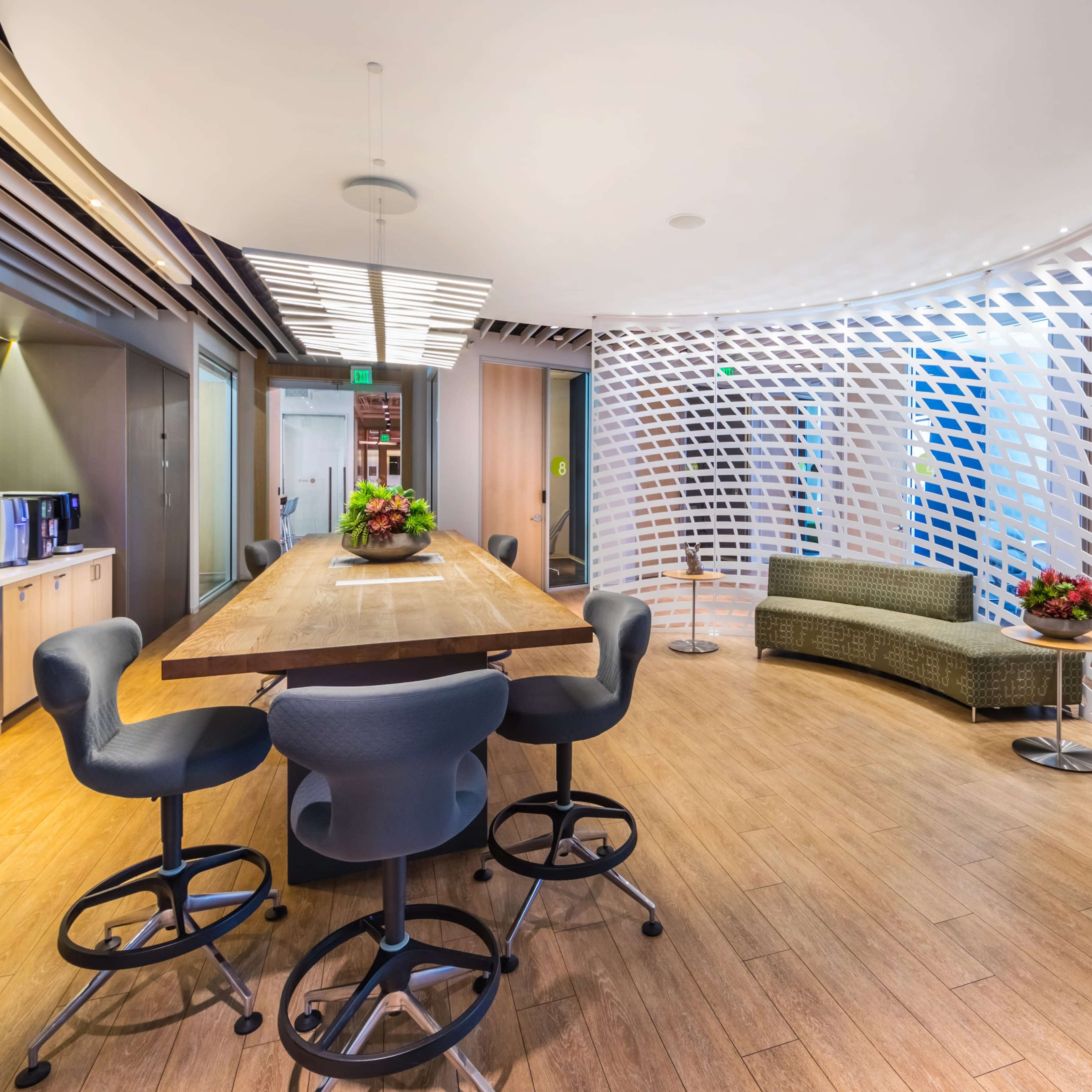 A modern office space features a large wooden table surrounded by high-backed chairs, with a decorative partition separating a lounge area.