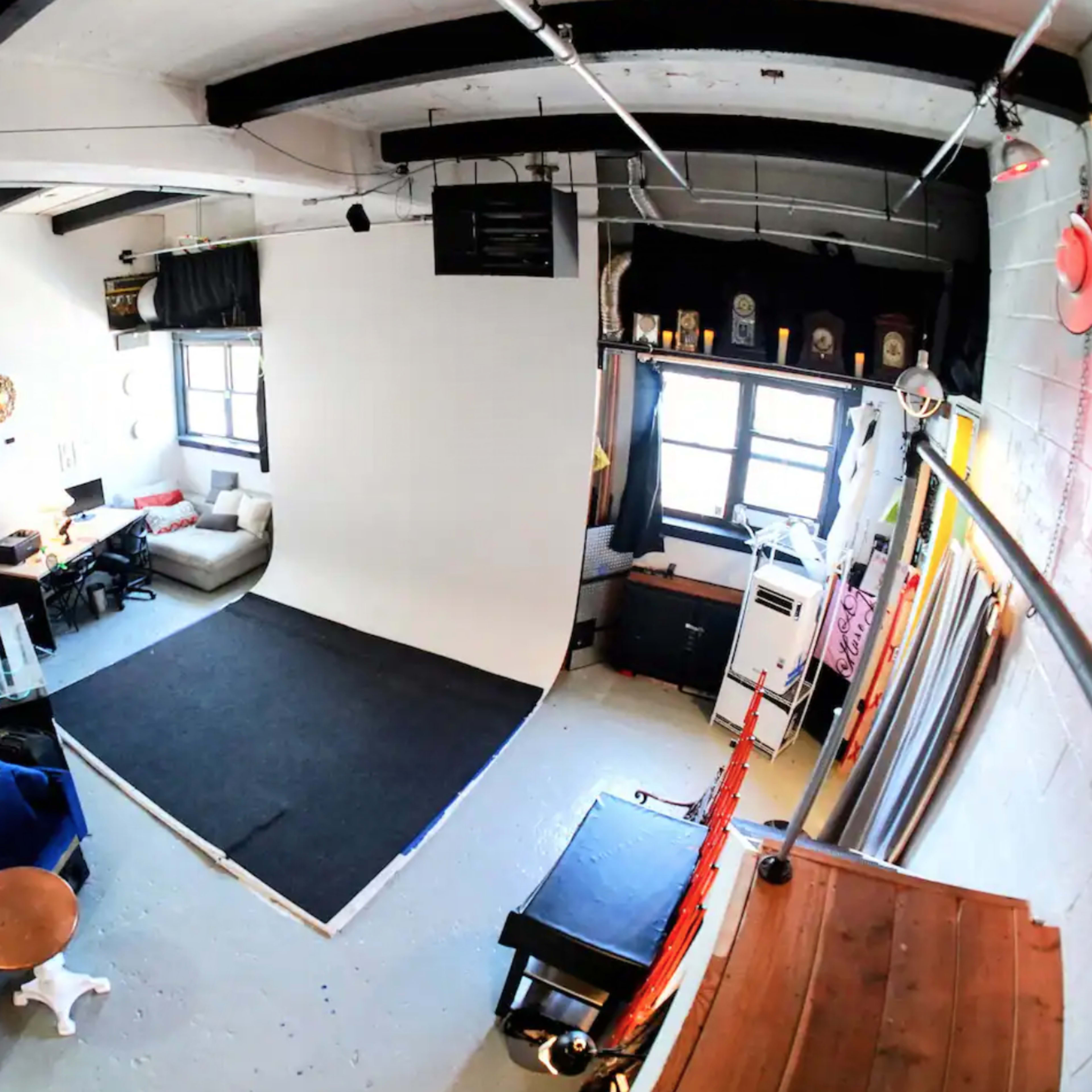 A spacious studio with exposed brick walls, a seating area with a blue couch, a photo backdrop, and various furniture pieces arranged around the space.