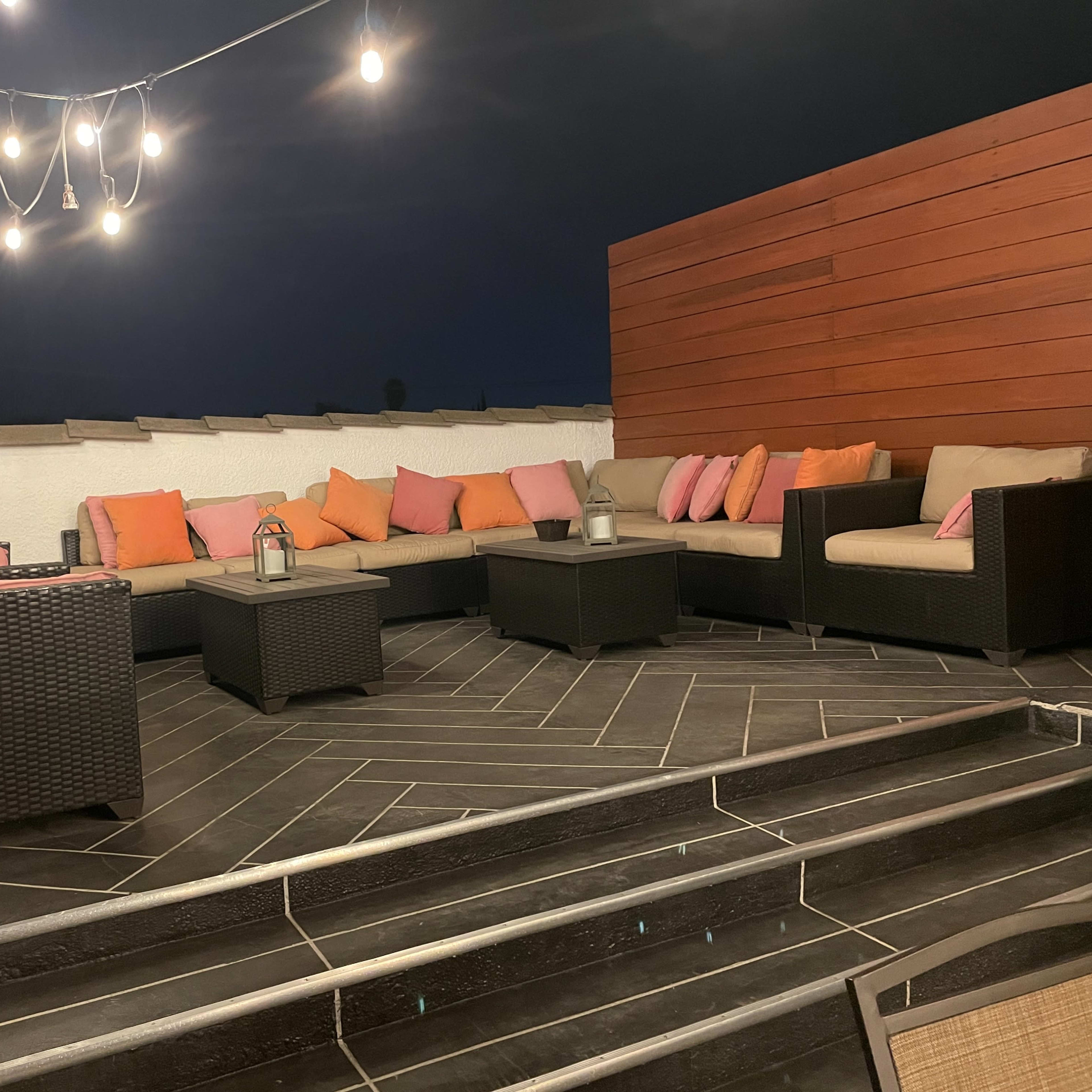 A rooftop seating area with a sectional couch adorned with colorful pillows, surrounded by string lights and wooden panels.