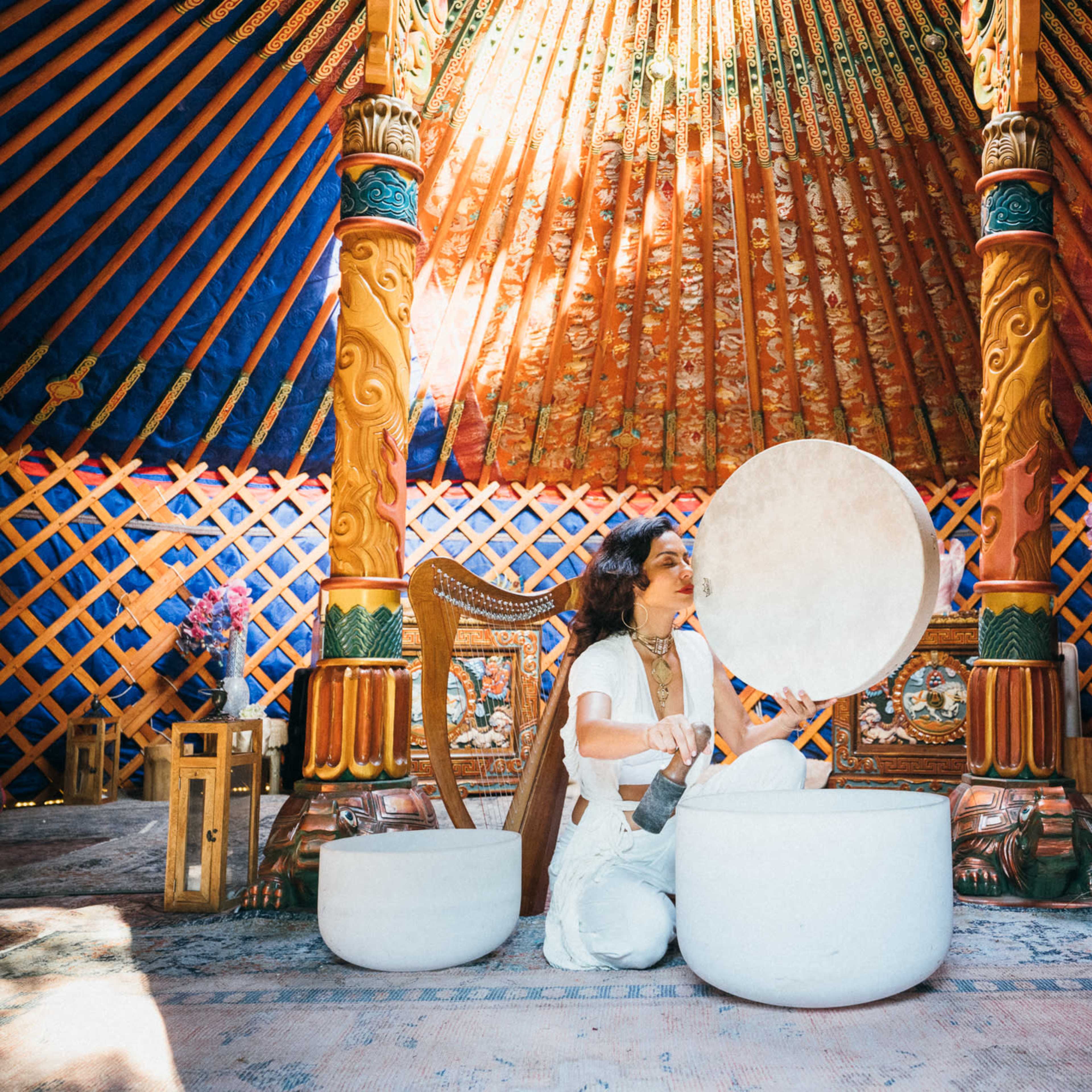 A person plays a large white singing bowl inside a colorful yurt adorned with intricate wooden details and vibrant patterns.