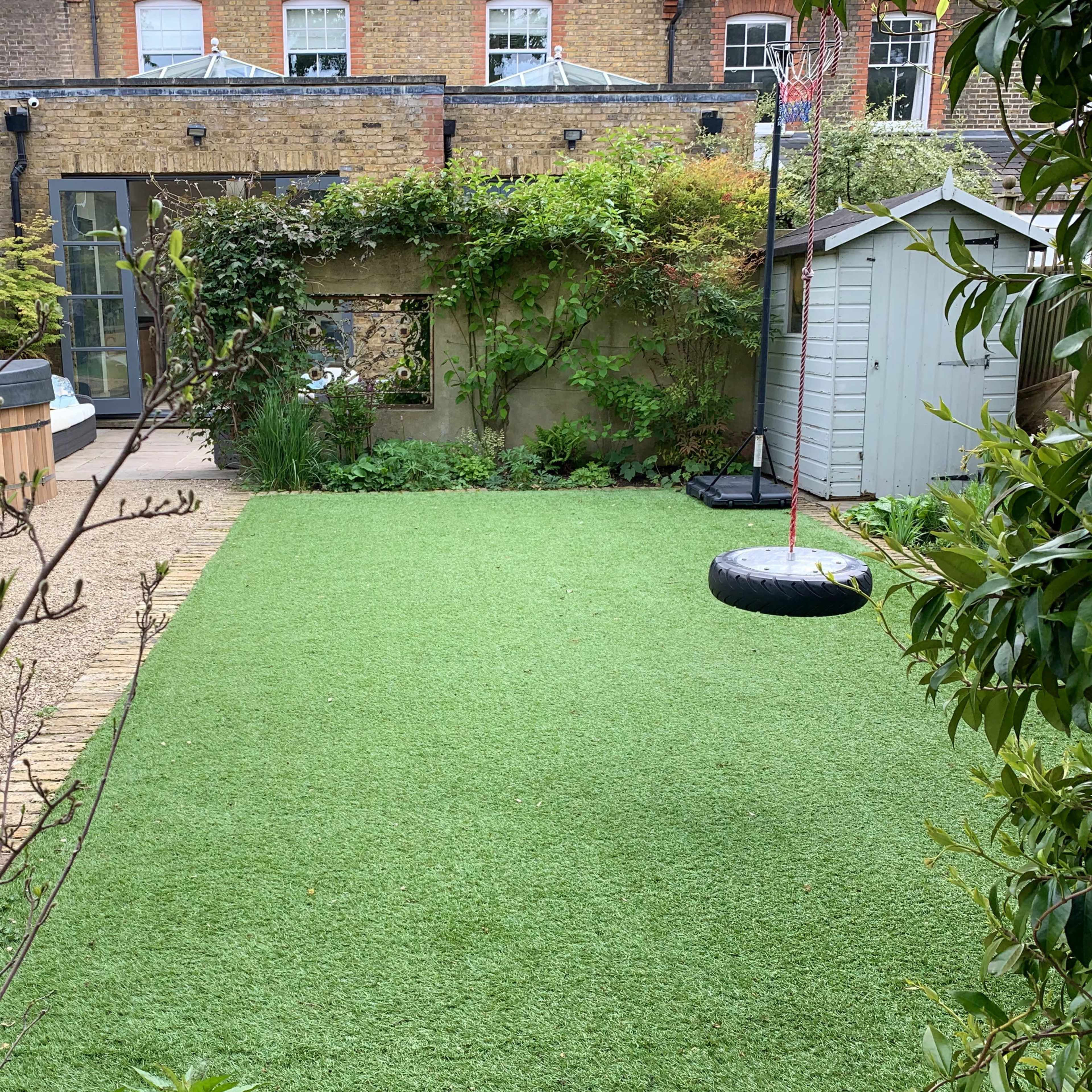 A well-maintained garden featuring a green synthetic lawn, a wooden hot tub, and a small shed, with a swing made from a tire hanging from a structure.