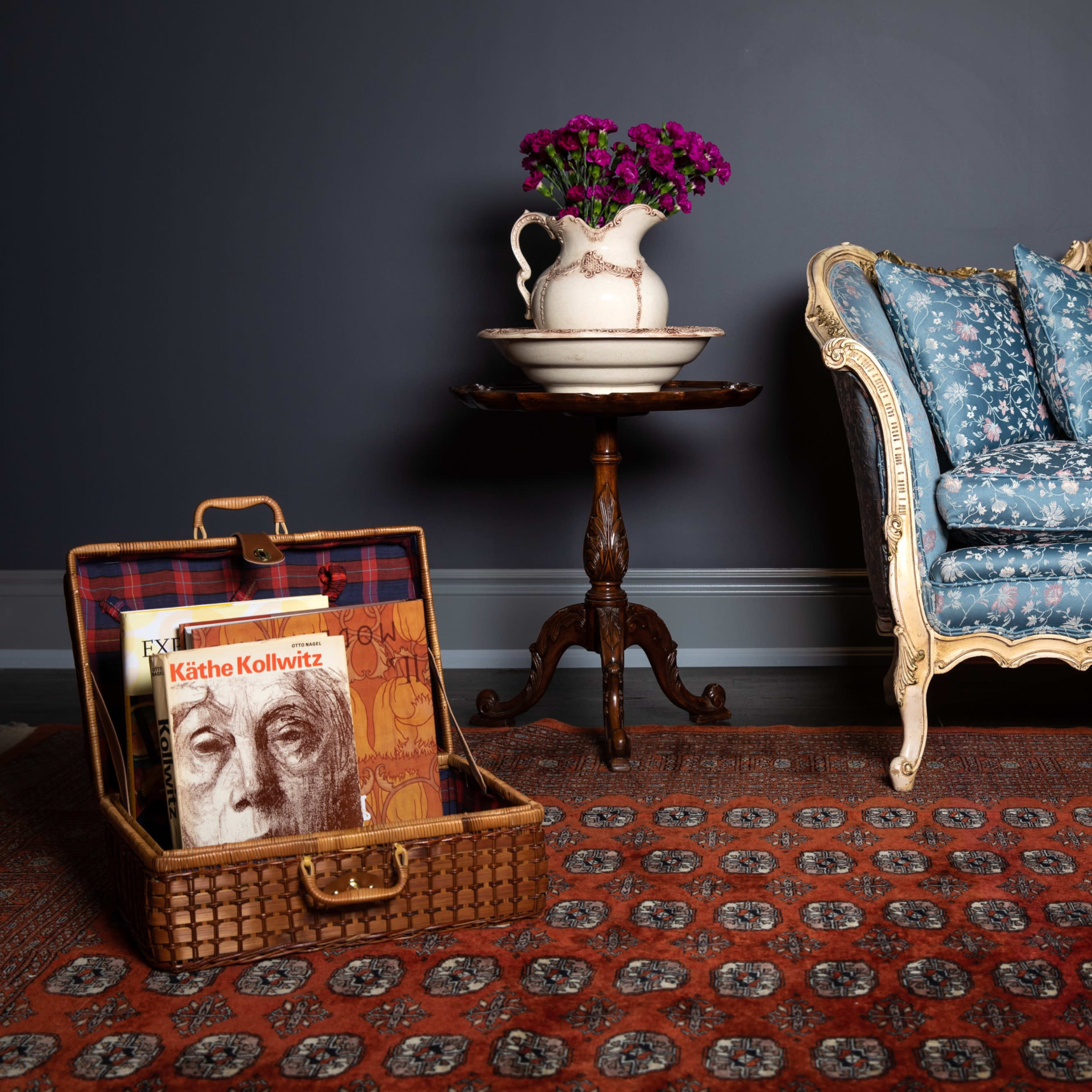 A vintage blue floral couch with decorative trim is positioned beside a small round table holding a vase of flowers, while a wicker basket containing a book rests on the patterned rug.