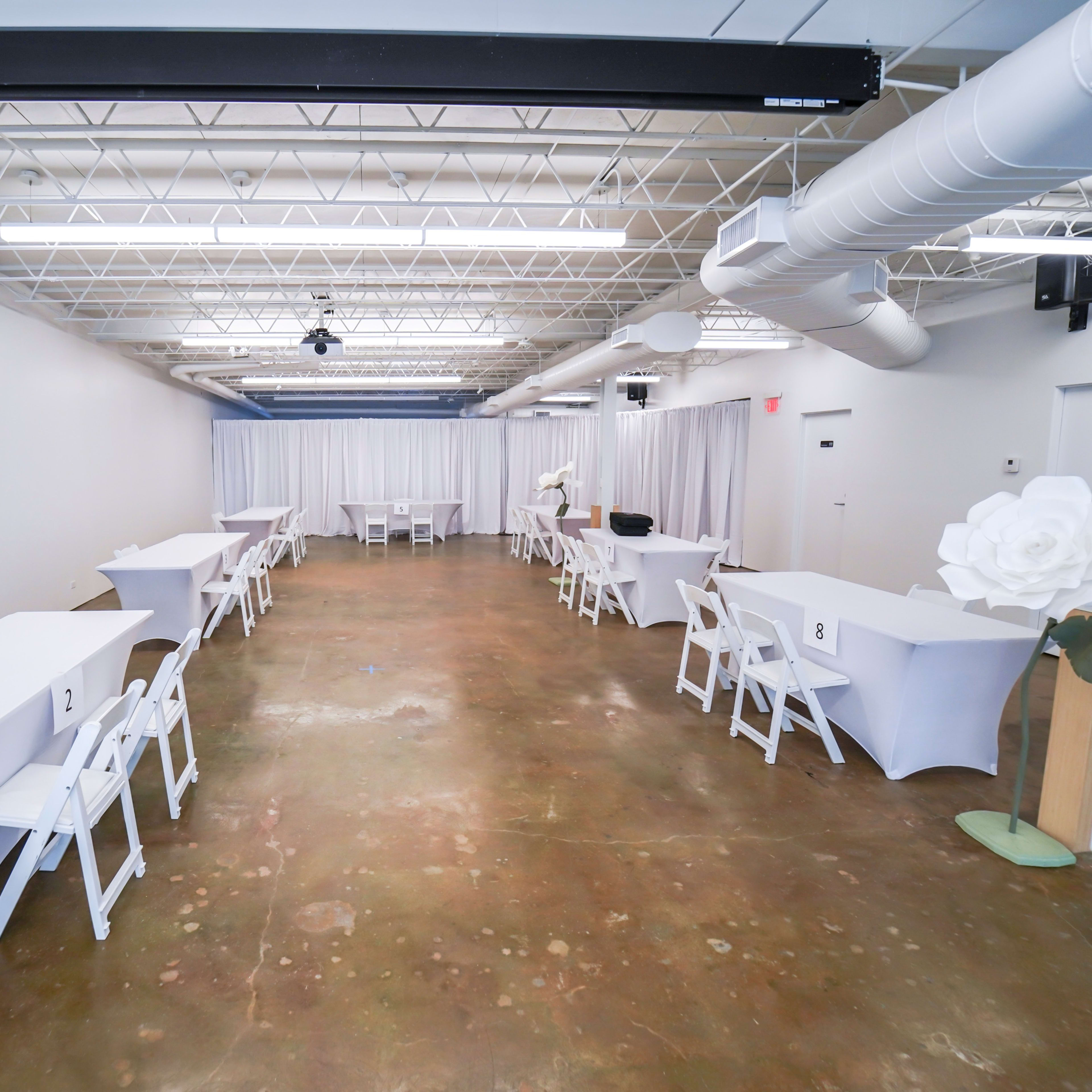 A spacious indoor venue features several white tables arranged with chairs and draped curtains at one end.