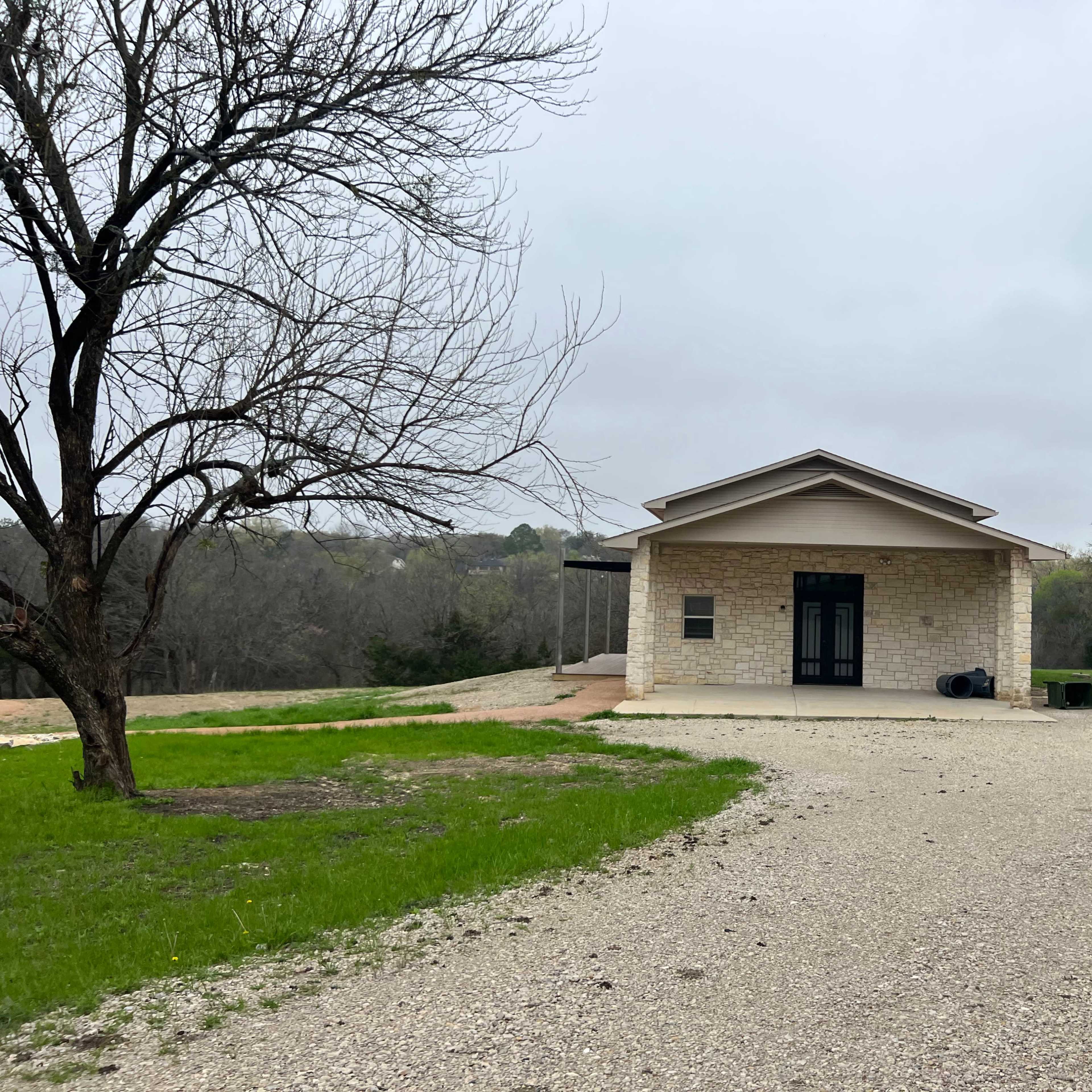 A single-story stone house is set on a gravel driveway surrounded by sparse trees and open land.