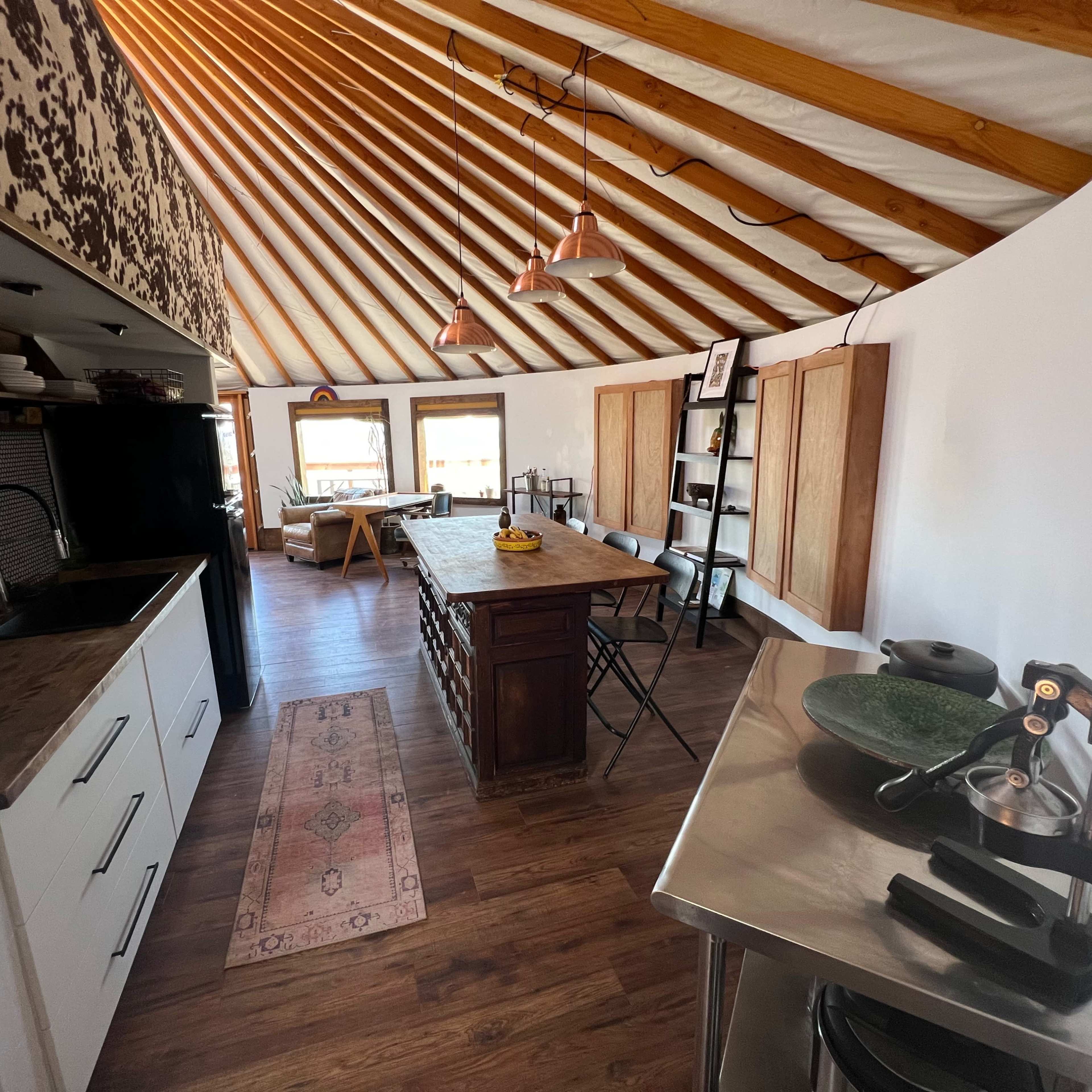 A modern kitchen inside a yurt, featuring a central island with a wooden countertop, a black refrigerator, and large windows allowing natural light.