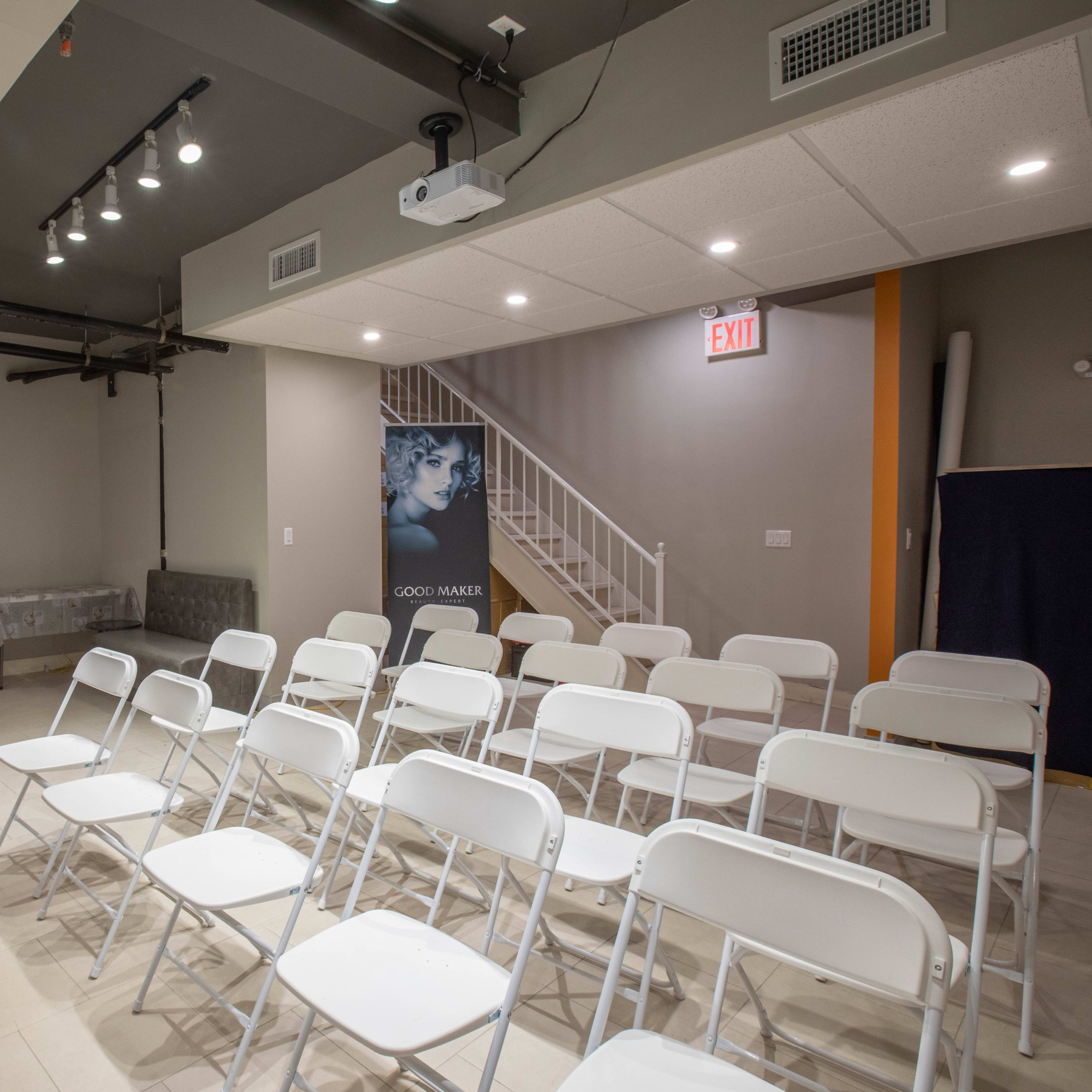 A minimalistic indoor space with white folding chairs arranged in rows facing a wall, near a gray couch and a staircase, with a promotional banner in one corner.
