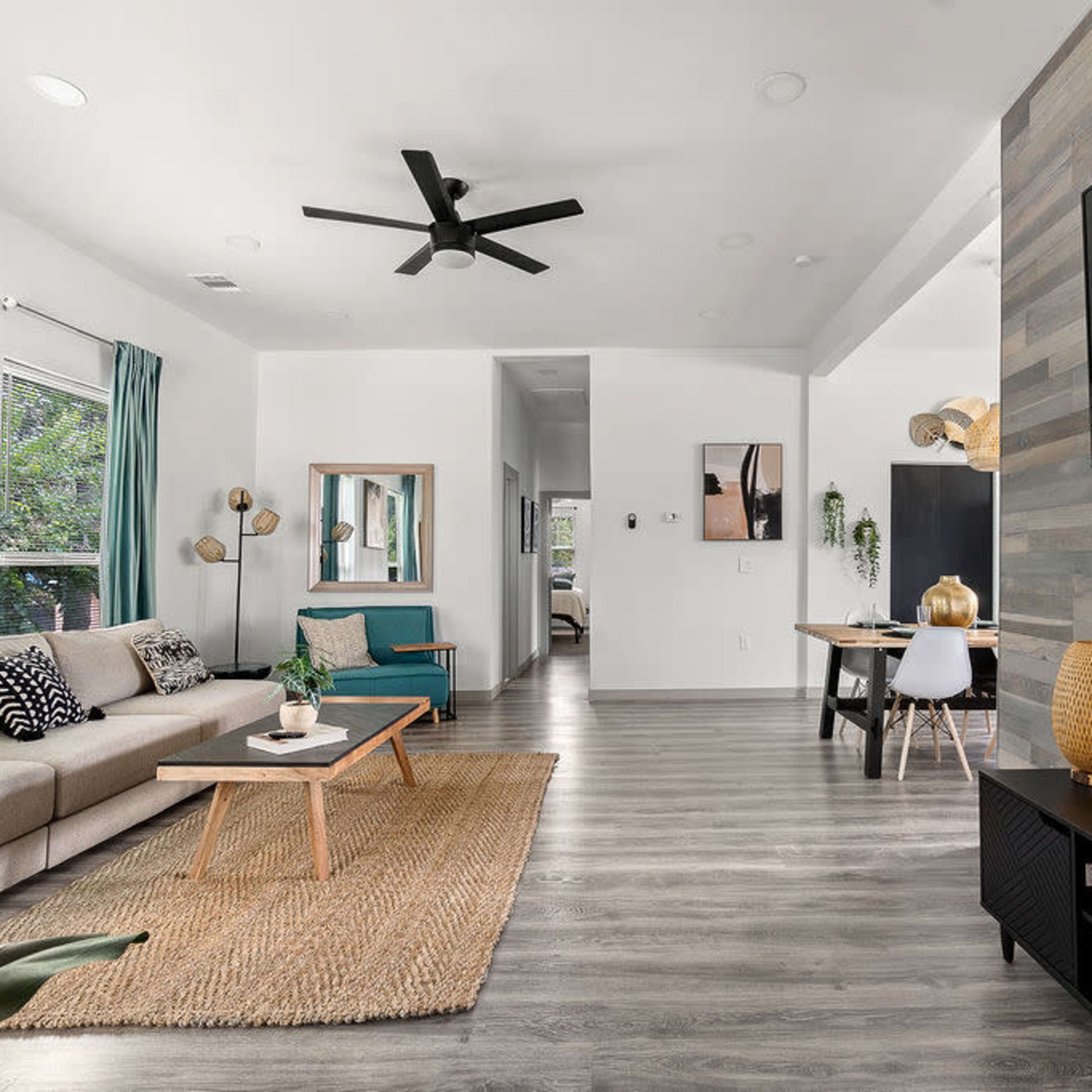 A modern living room with a gray sofa, a teal armchair, and a wooden dining area in the background, featuring a large flat-screen TV mounted on a textured wall.