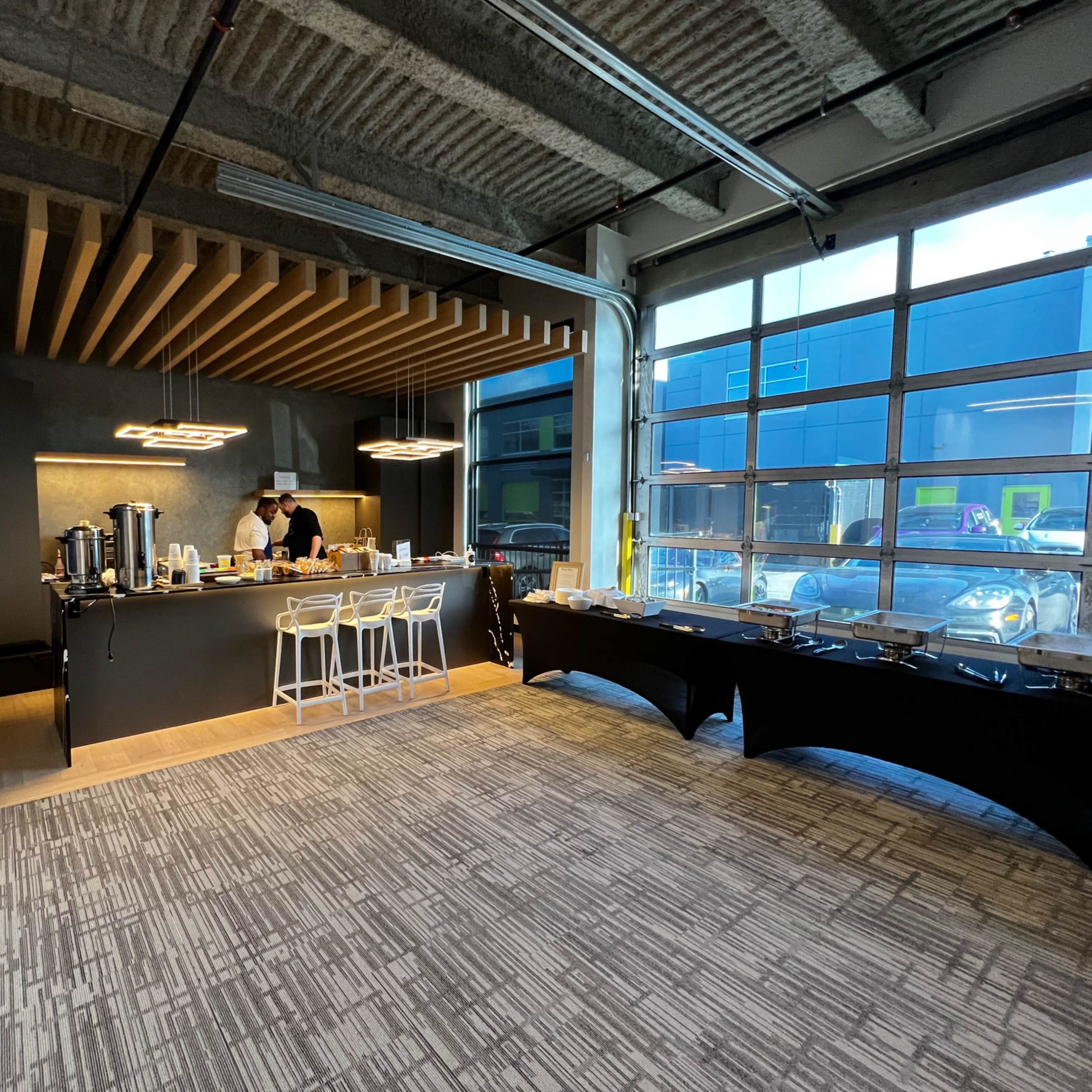 A modern event space features a sleek bar area with a countertop, a person preparing food, and tables set up for catering.