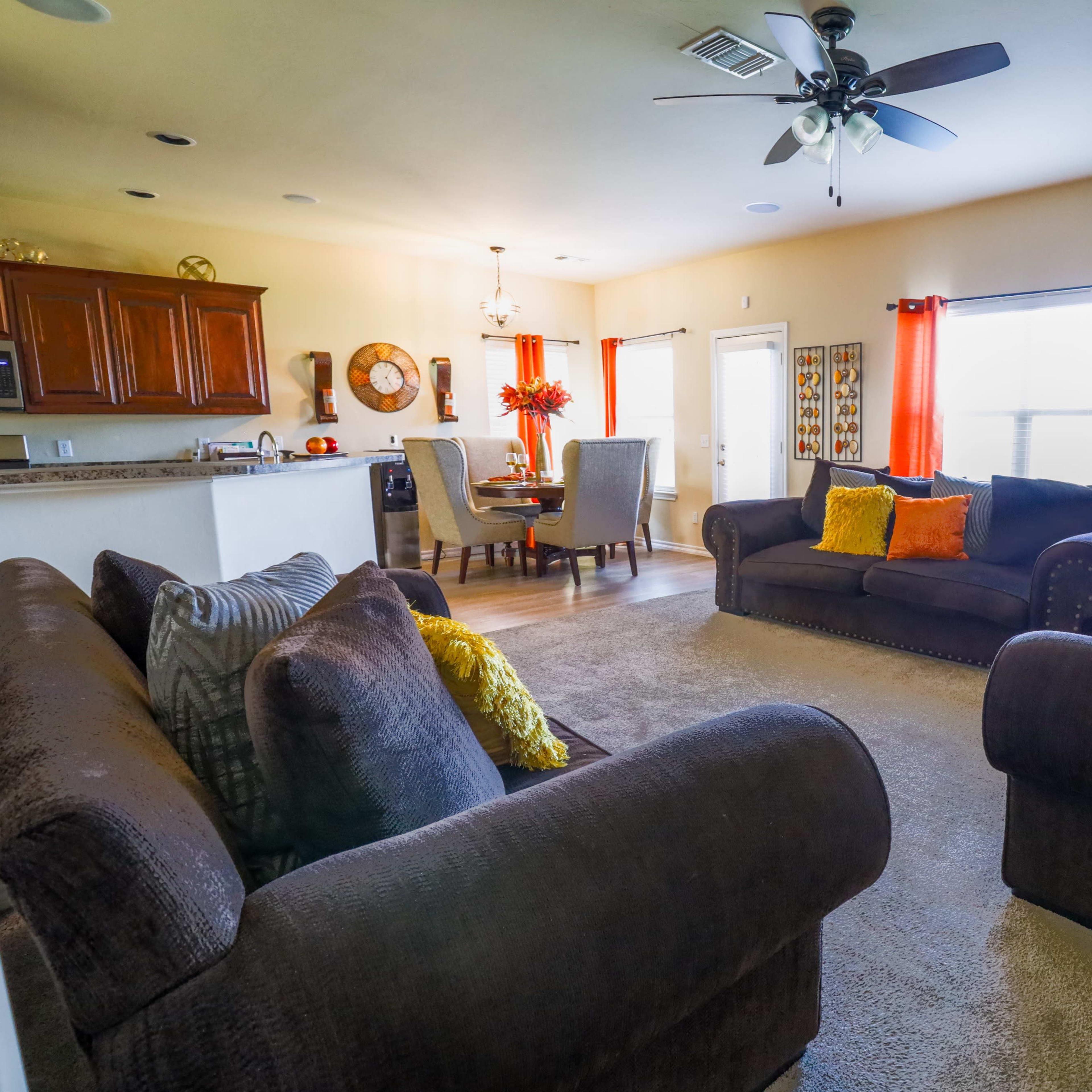 A cozy living room with two dark sofas facing each other, a round dining table with chairs, a fireplace, and large windows with orange curtains.