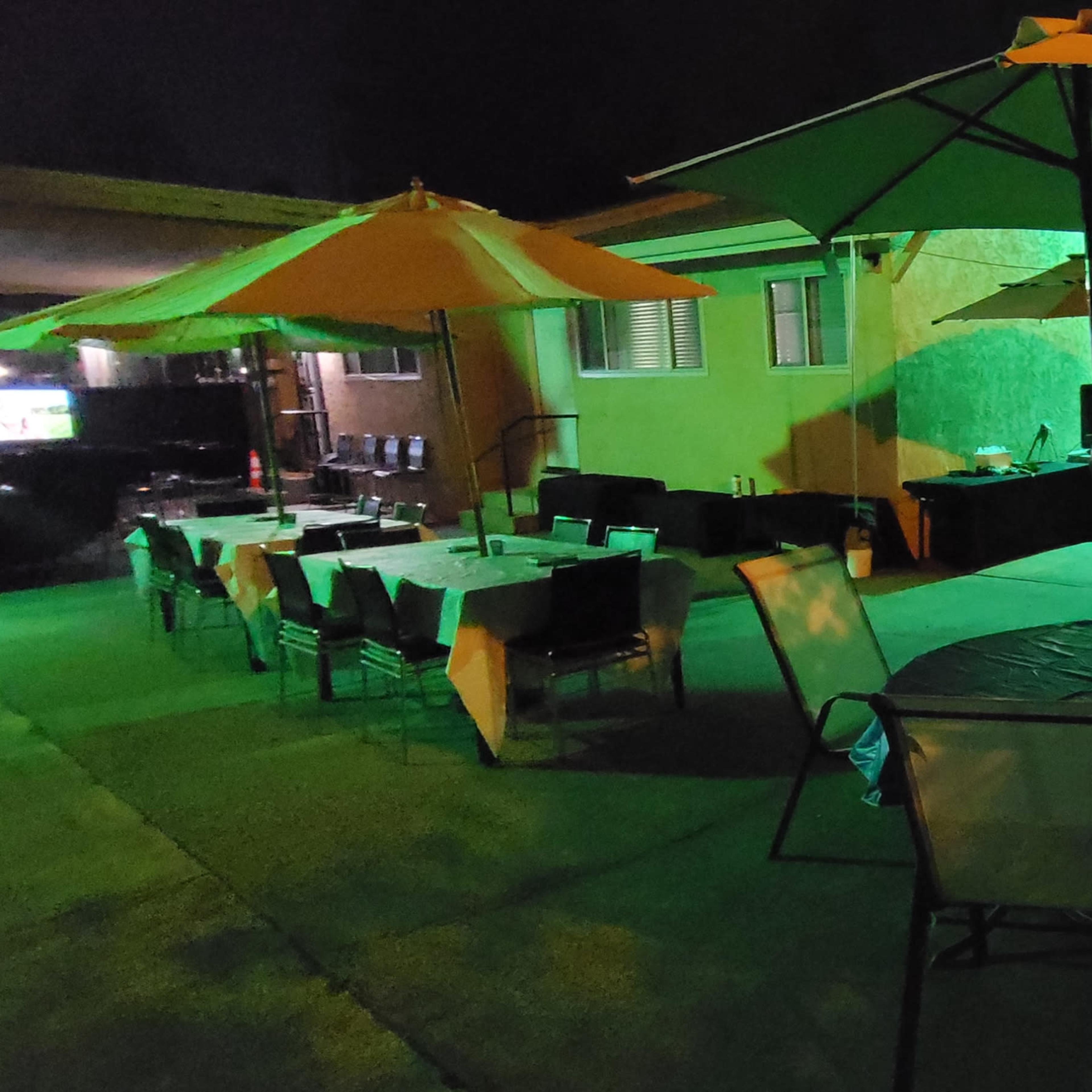 A nighttime outdoor gathering features tables and chairs under yellow umbrellas, with green lighting illuminating the area.