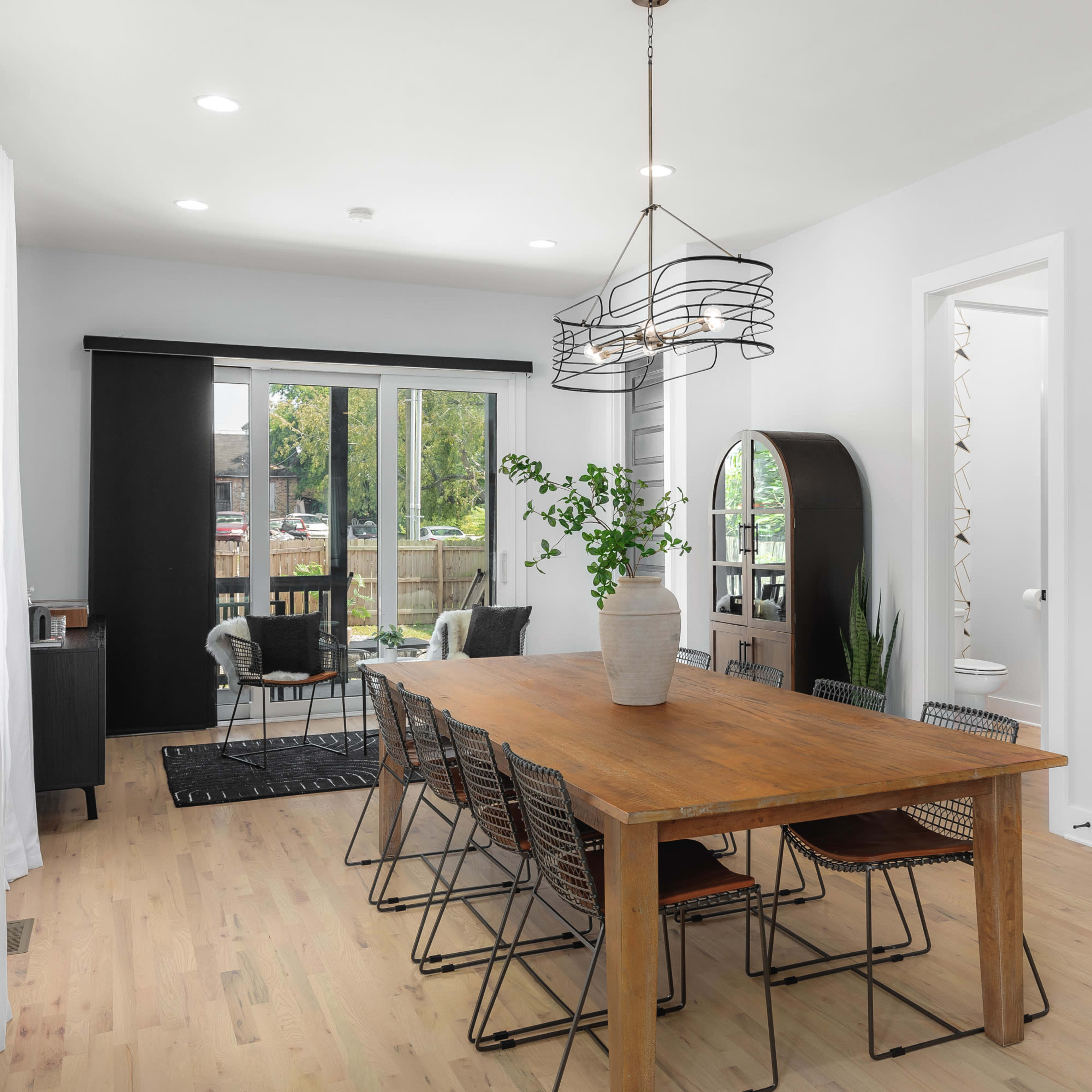 A spacious dining area features a large wooden table surrounded by metal chairs, with a modern light fixture above and sliding glass doors leading to an outdoor space.