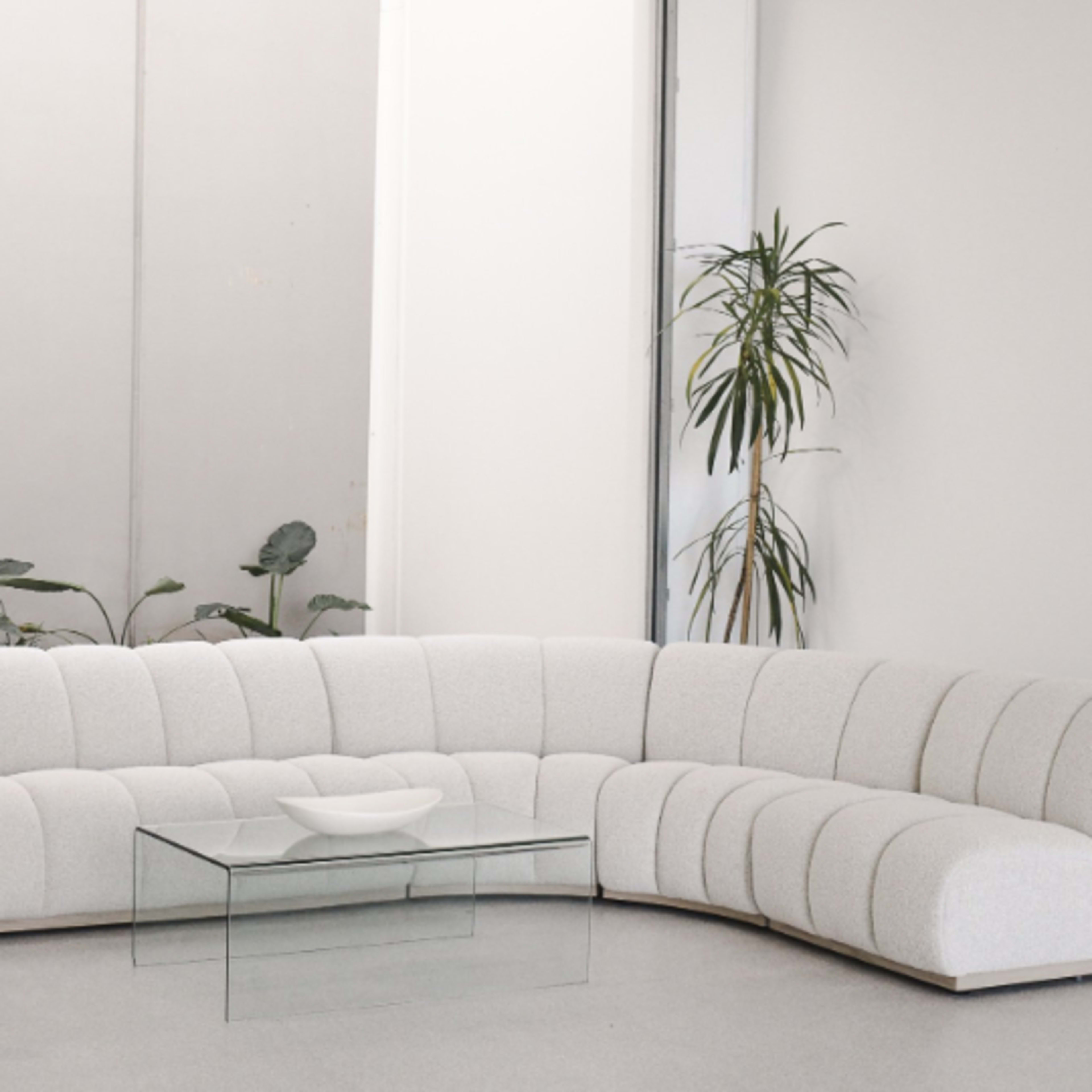 A curvy, light-colored sectional sofa with tufted upholstery is positioned next to a glass coffee table in a minimalist room with indoor plants.