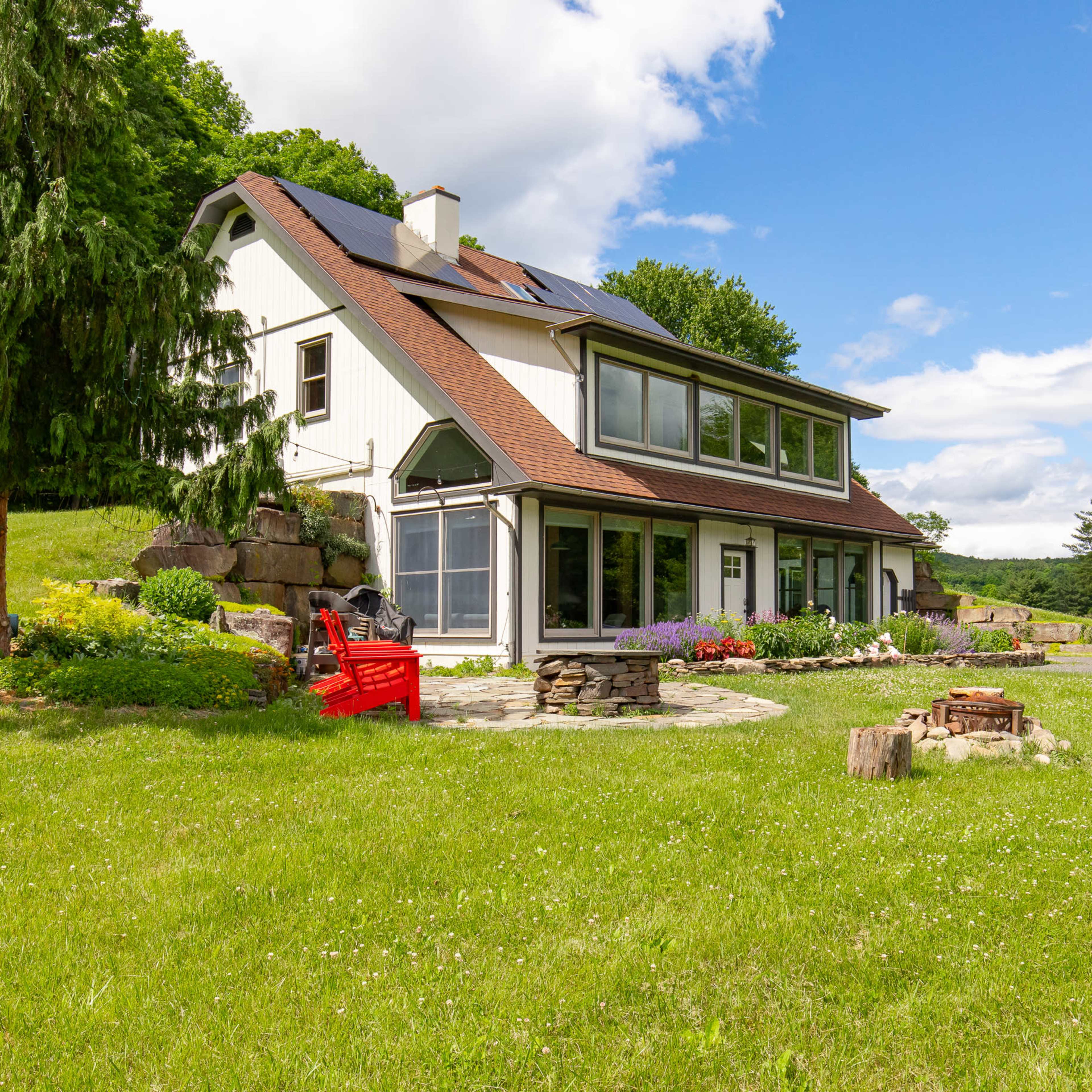 A modern, two-story house is situated on a grassy lawn with a stone patio and fire pit, surrounded by trees and landscaped gardens.