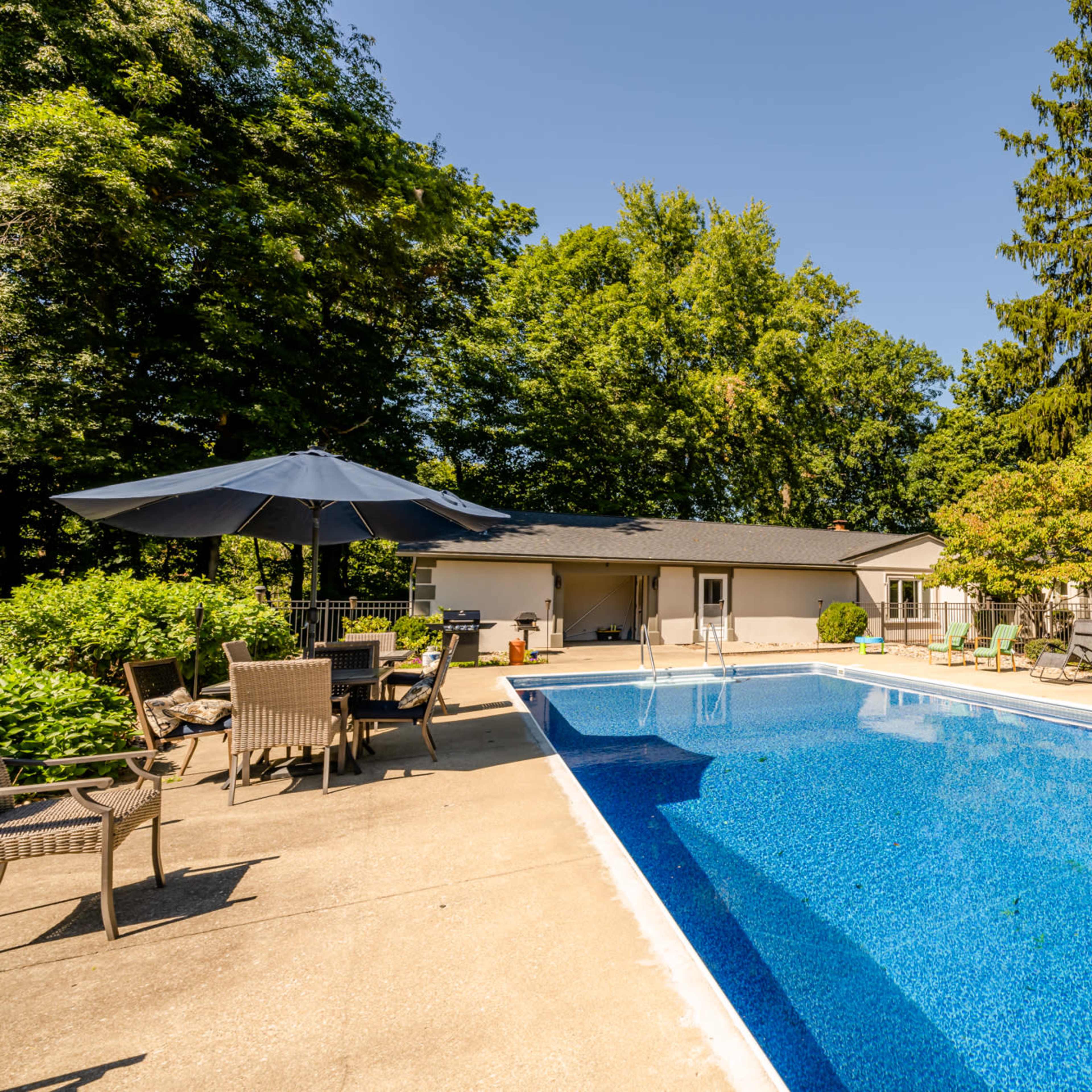 A clear swimming pool surrounded by patio furniture and a wooded area in the background.