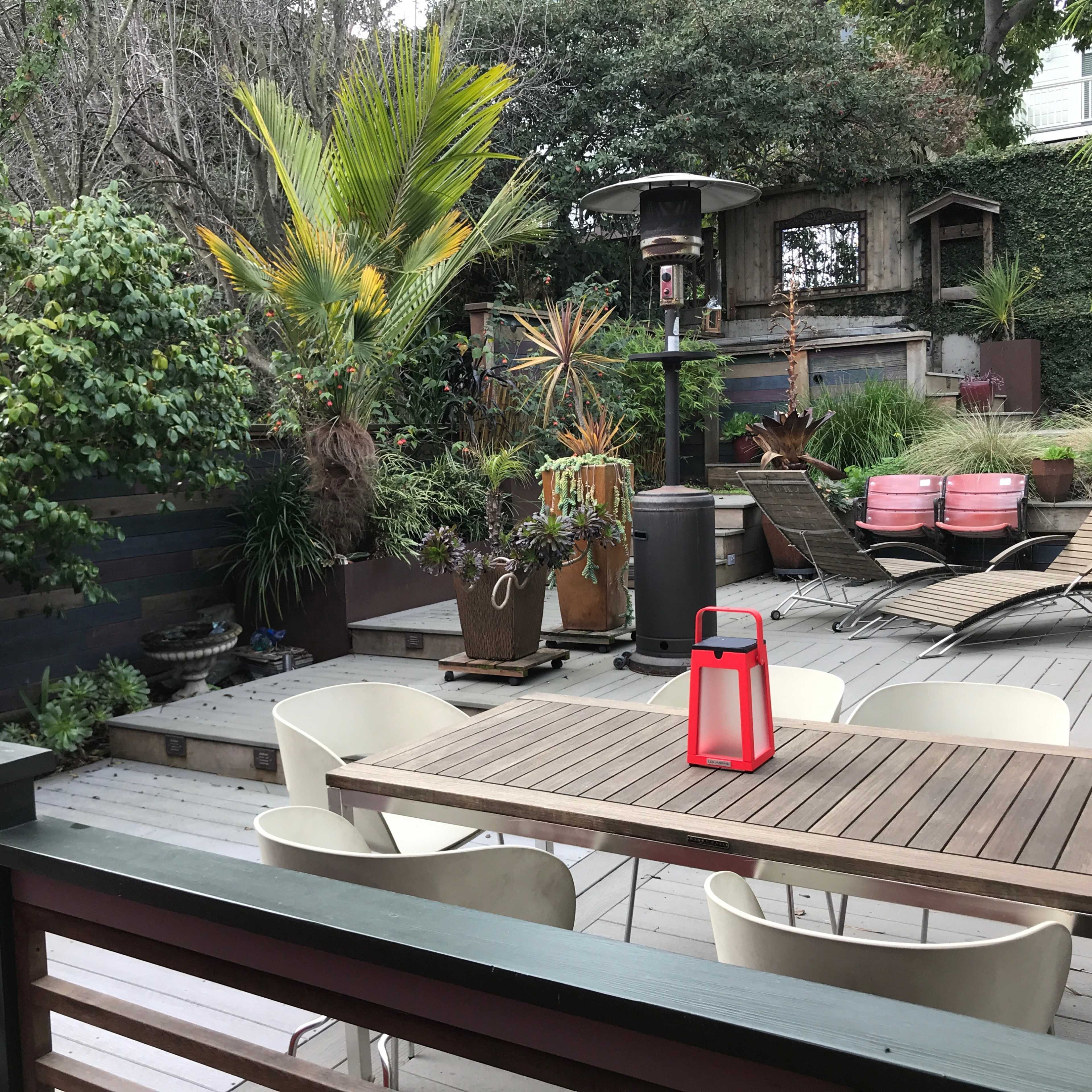 A wooden deck with outdoor seating, a table, and a patio heater surrounded by lush greenery and decorative plants.
