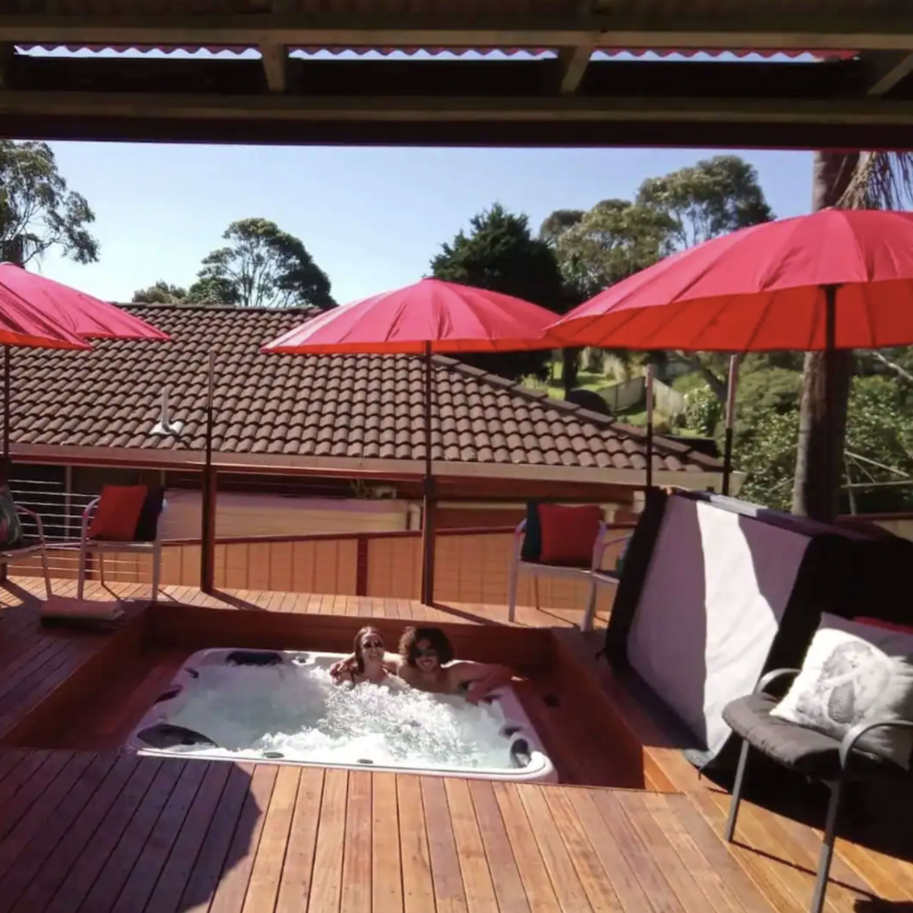 A wooden deck features a hot tub surrounded by red umbrellas and overlooks a house with a tiled roof.