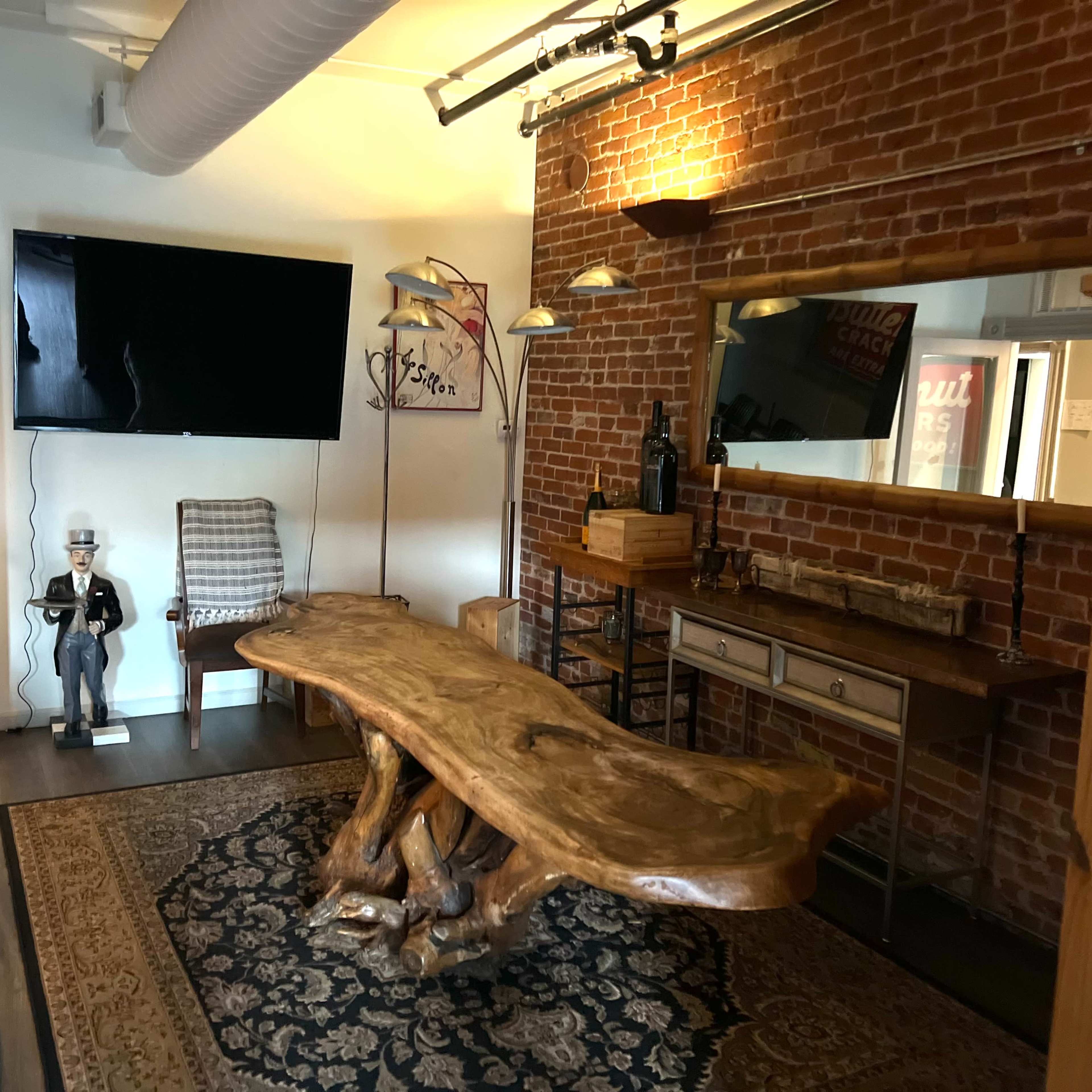 A room with a large wooden table, a vintage-style chair, a flat-screen TV mounted on a brick wall, and a mirror above a console table.