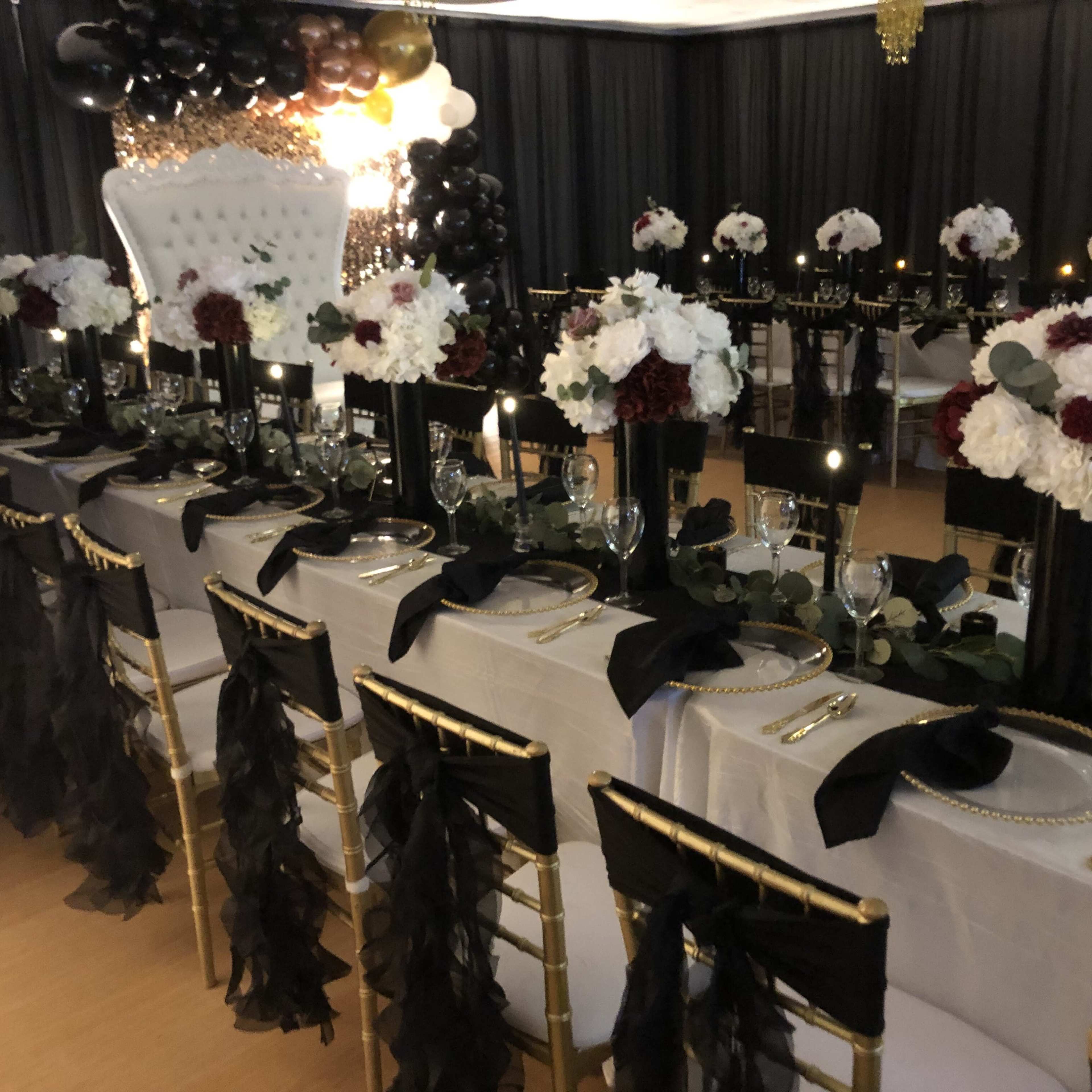 A beautifully arranged banquet long table with elegant black and white decor, featuring floral centerpieces, black chair sashes, and golden accents.