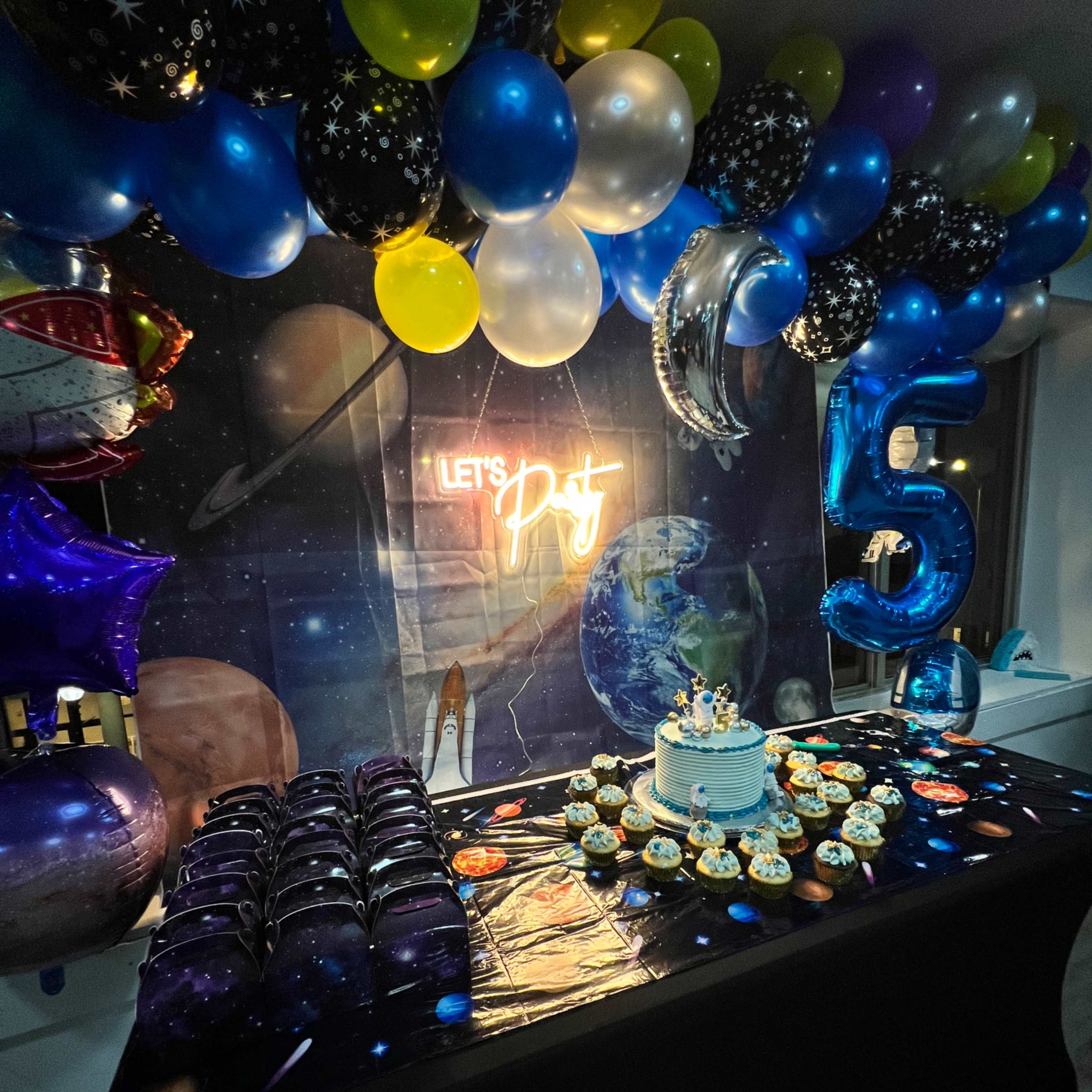 A birthday party setup with a table covered in a space-themed tablecloth, decorated with a birthday cake, cupcakes, and balloons, alongside a glowing sign that reads "LET'S PARTY."