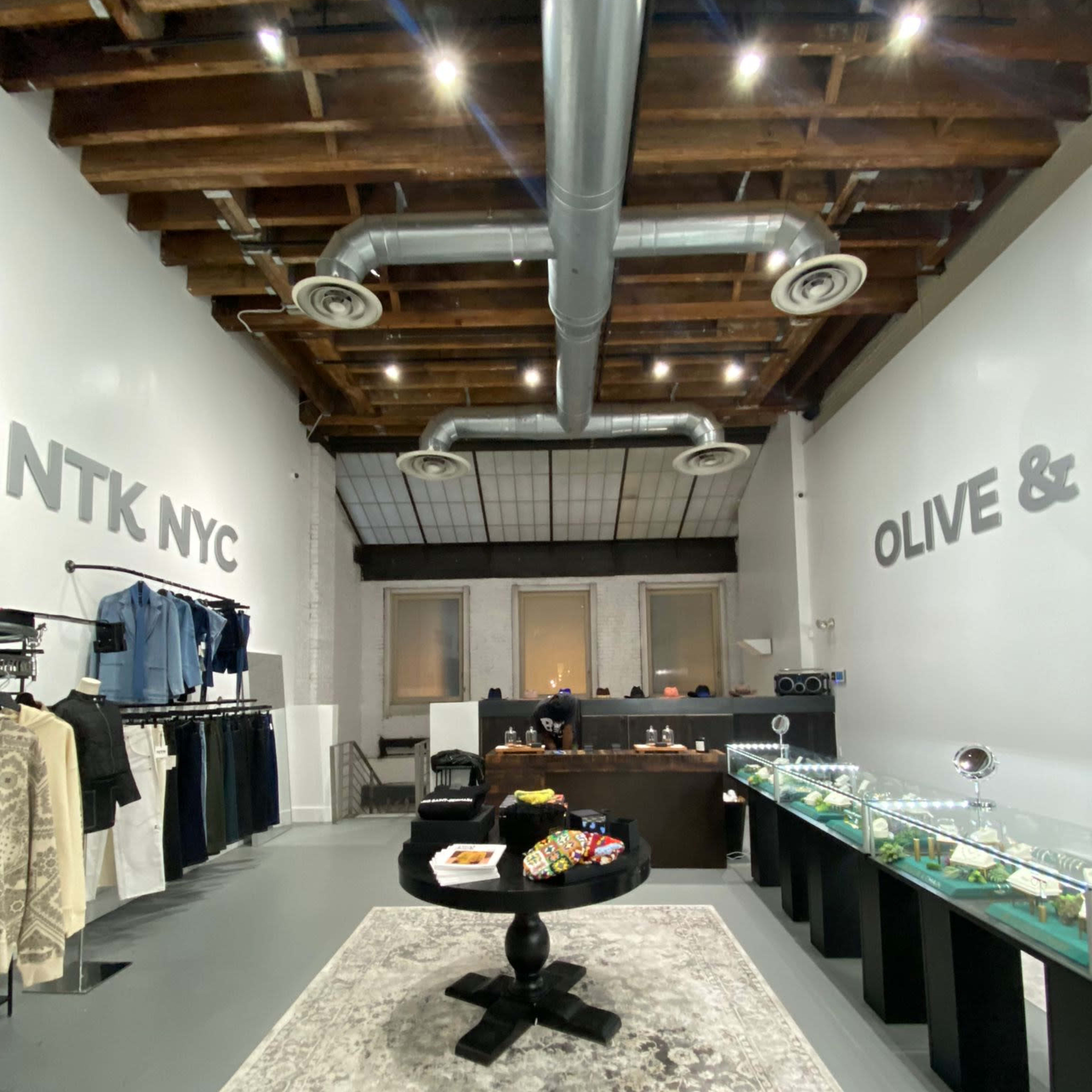 A modern retail space with clothing racks on the left, a display counter with glass showcases in the center, and large text on the walls reading "NTK NYC" and "OLIVE &".