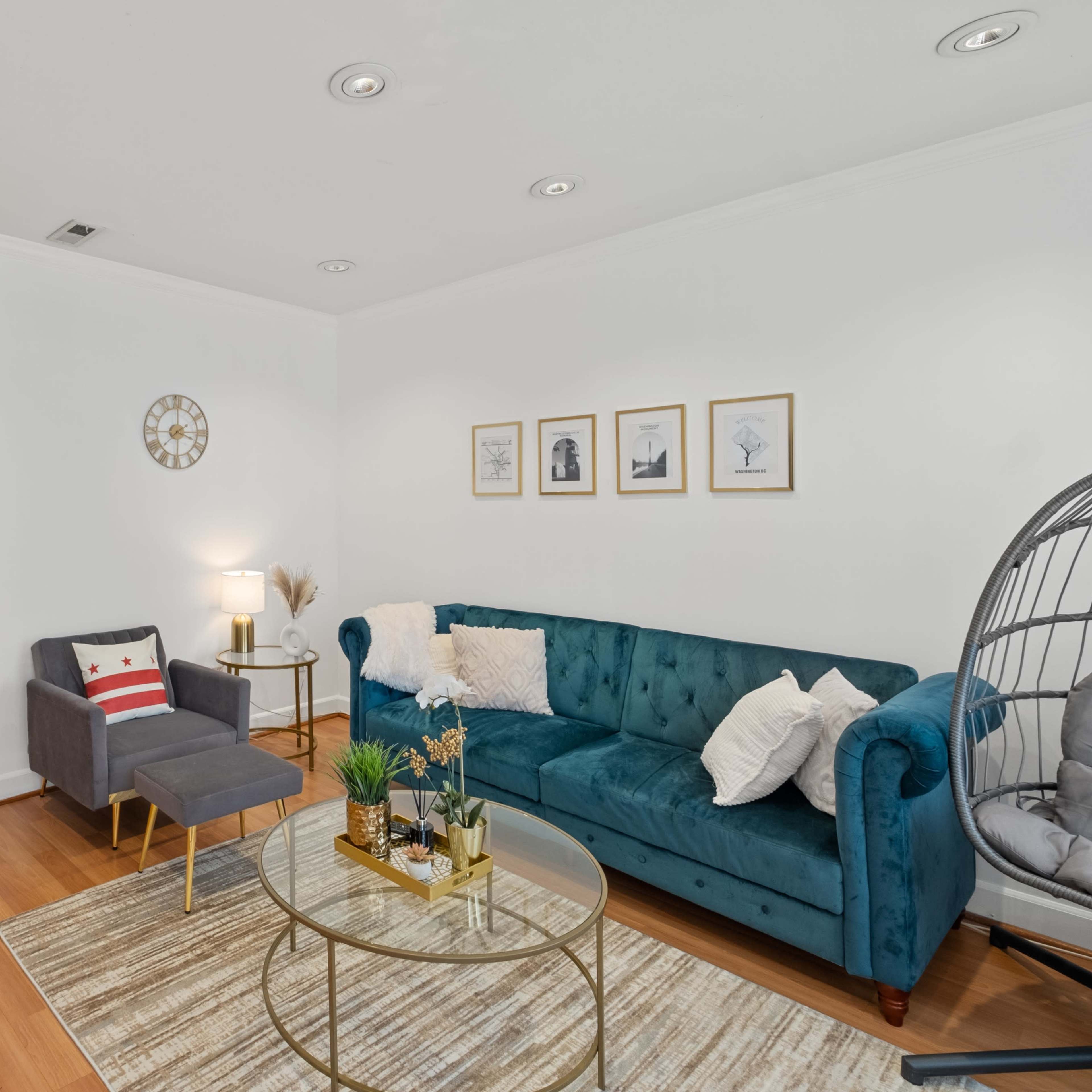 The living room features a teal sofa, a round coffee table, and framed artwork on the white walls, with a kitchen visible in the background.