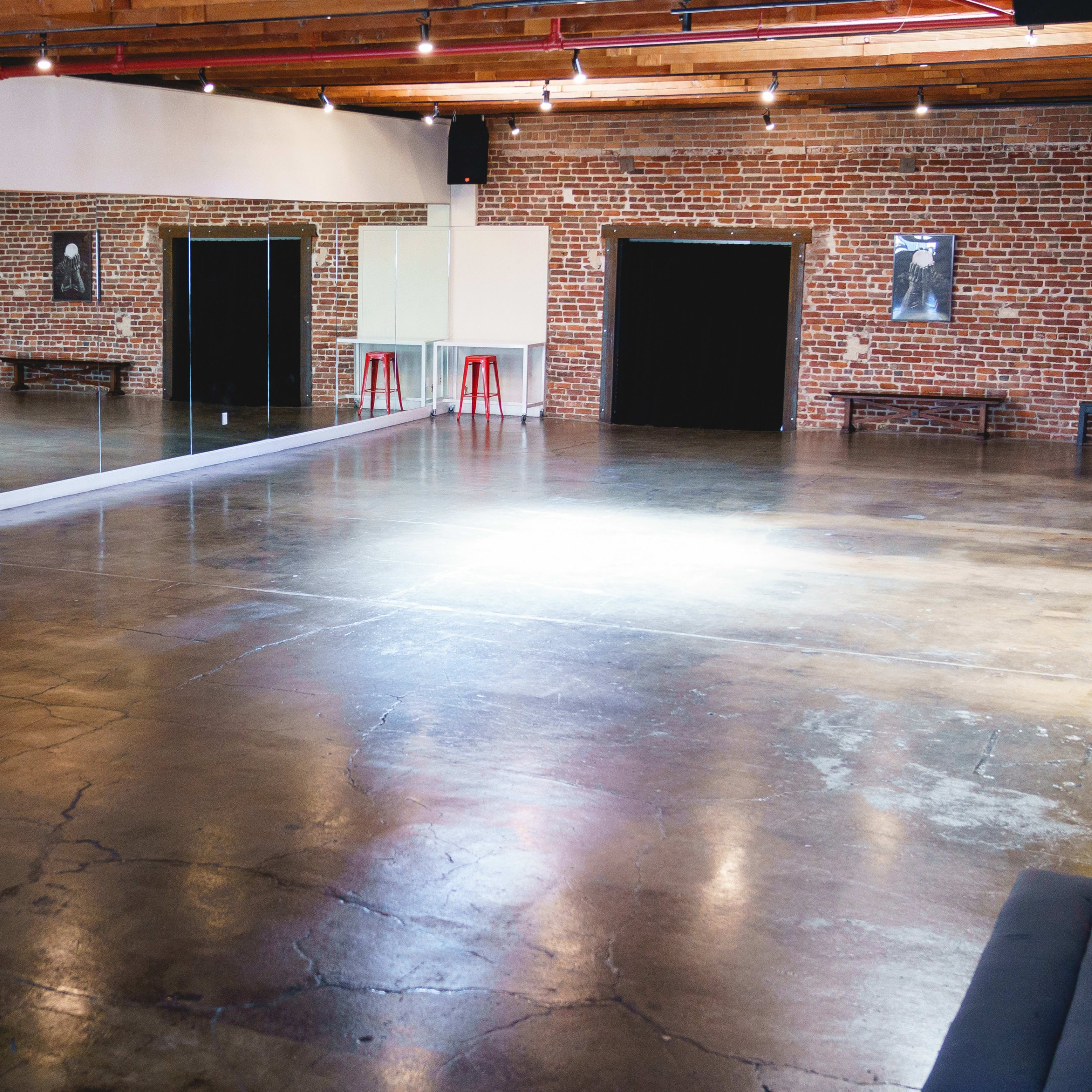 An empty dance studio with exposed brick walls, a large mirror, and polished concrete flooring.