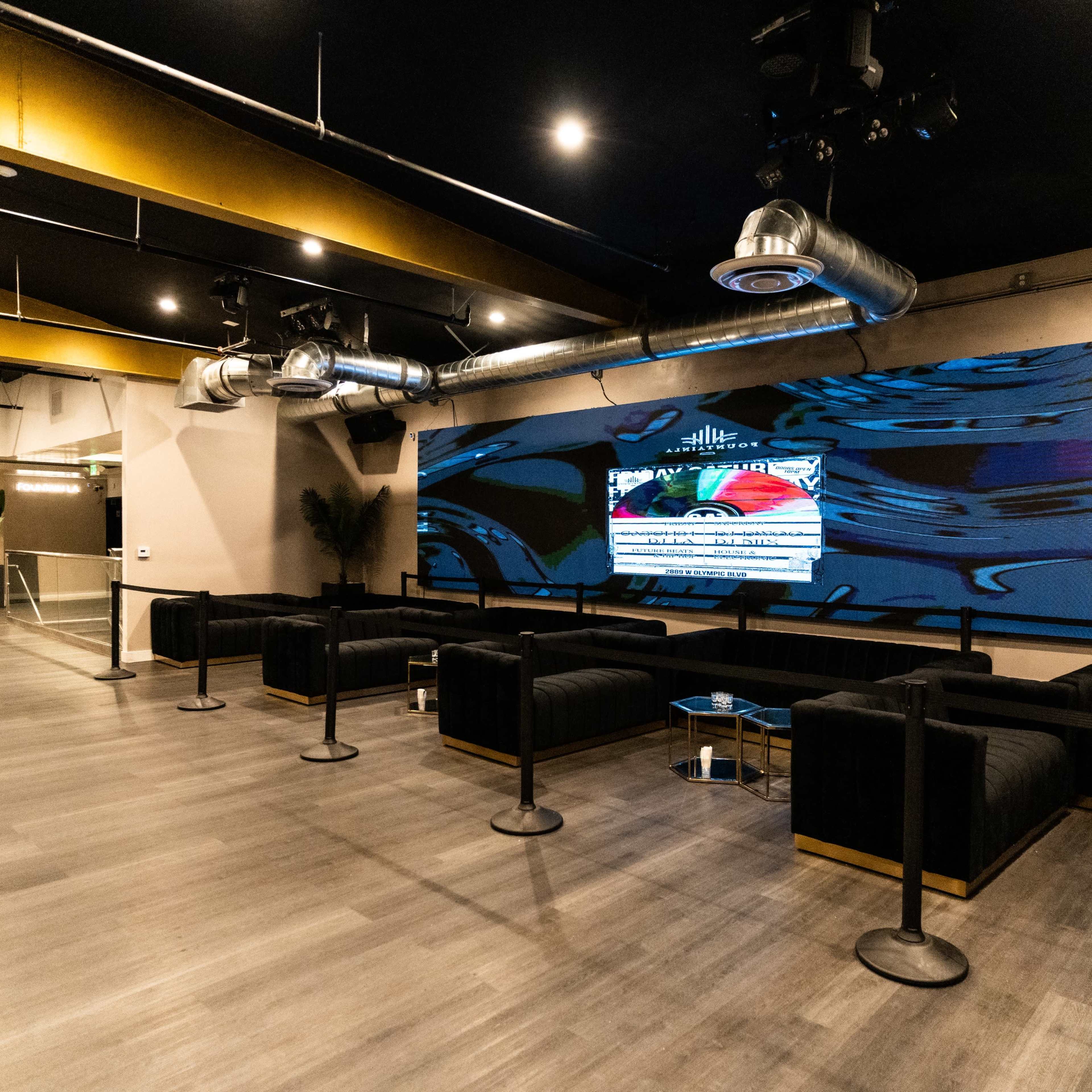 A modern lounge area featuring black seating, a large screen displaying visuals on a wall, and an open layout with a wooden floor.