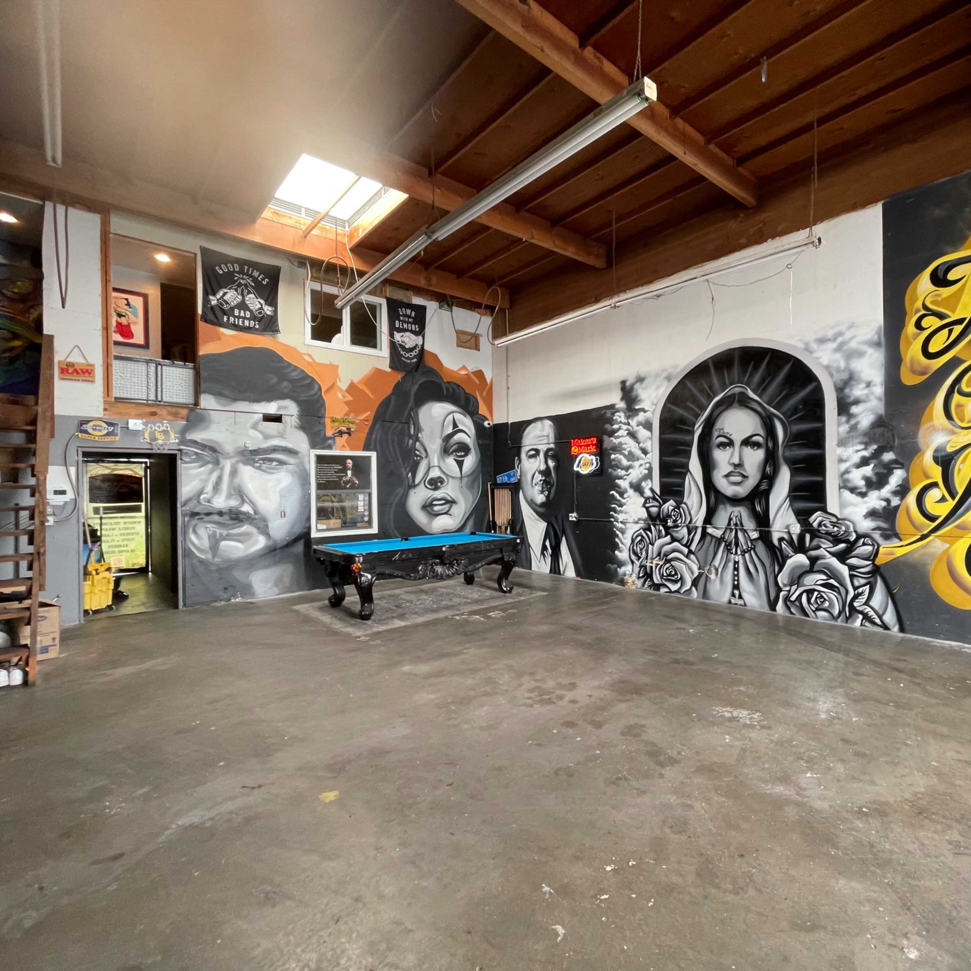An industrial-style room featuring large, intricate murals on the walls, a pool table in the center, and shelves with various items along one side.