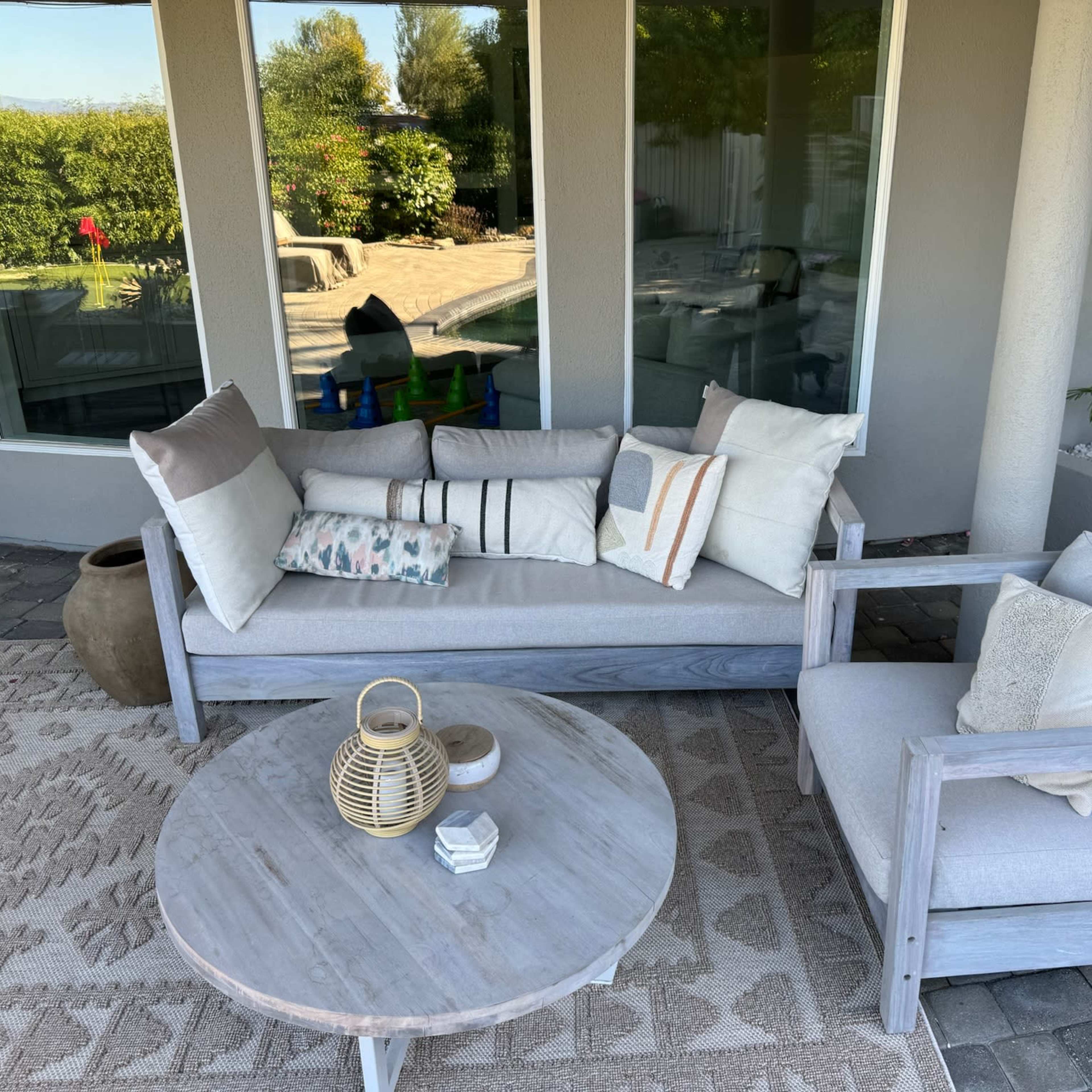 A modern outdoor seating arrangement features a light gray sofa and armchair, accompanied by a round coffee table with a lantern, set against a backdrop of large windows and a landscaped yard.