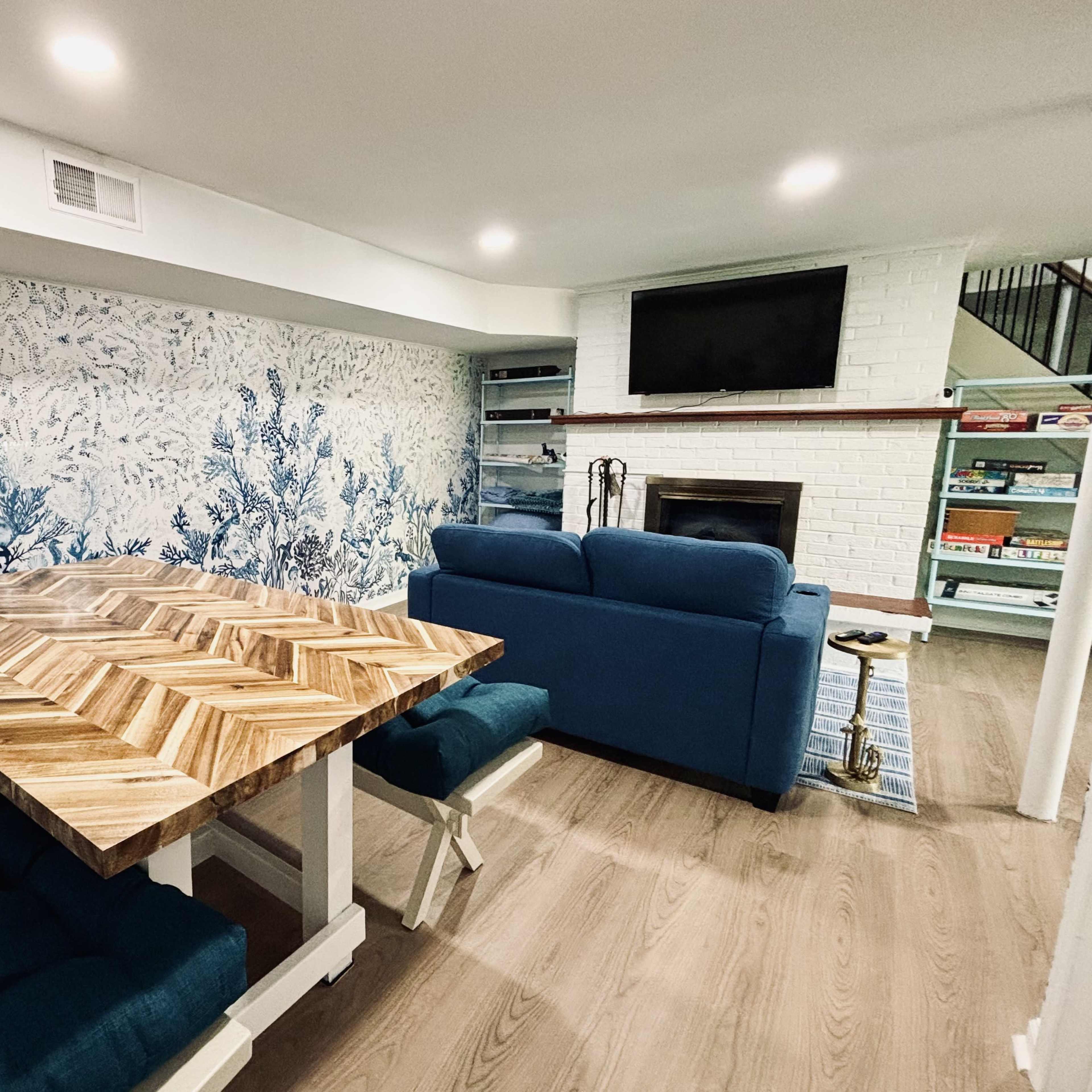 A living space features a patterned blue wallpaper, a wooden dining table, a blue couch, and a fireplace with a television mounted above it.