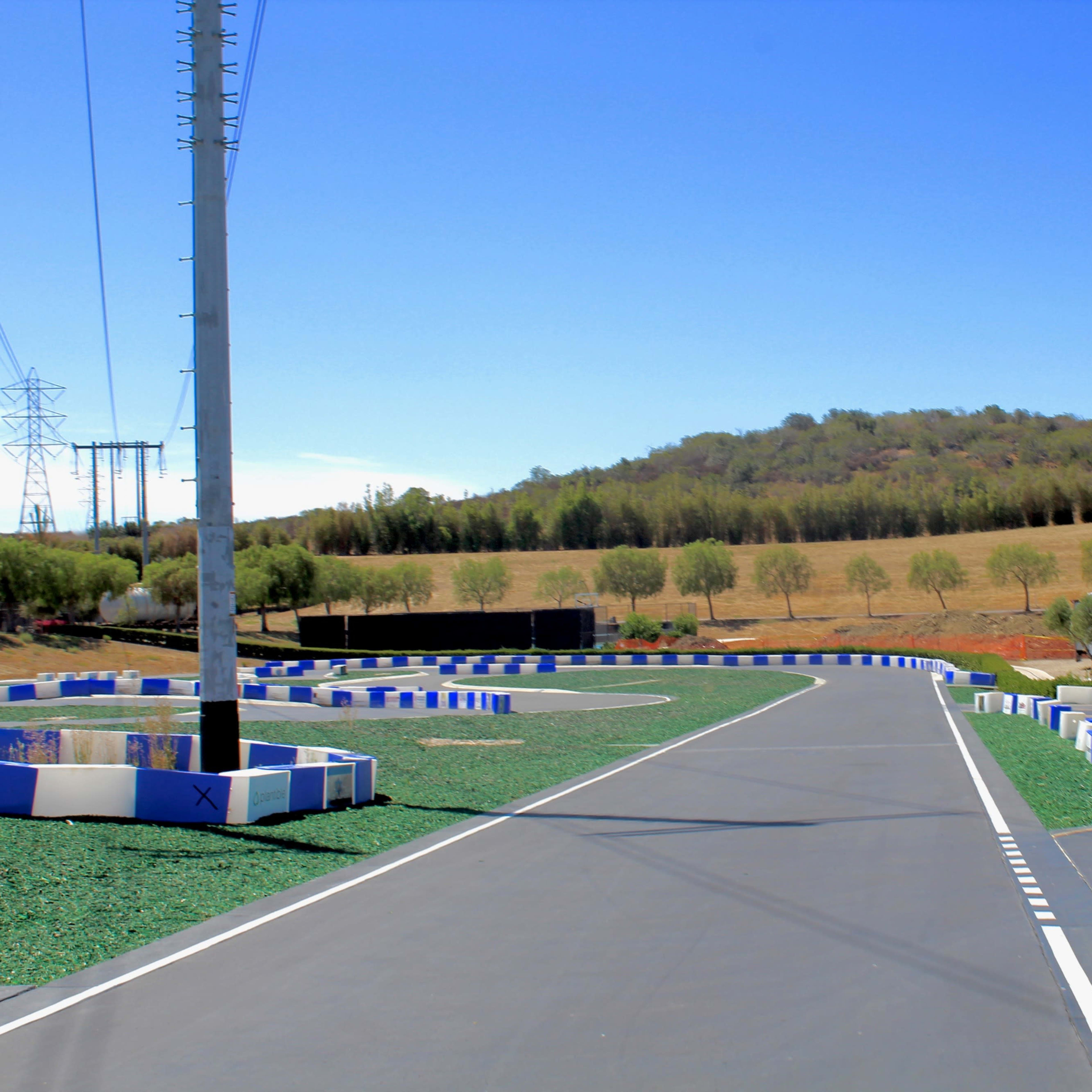 A racetrack with a winding asphalt path bordered by blue and white barriers, set against a backdrop of green hills and clear blue skies.