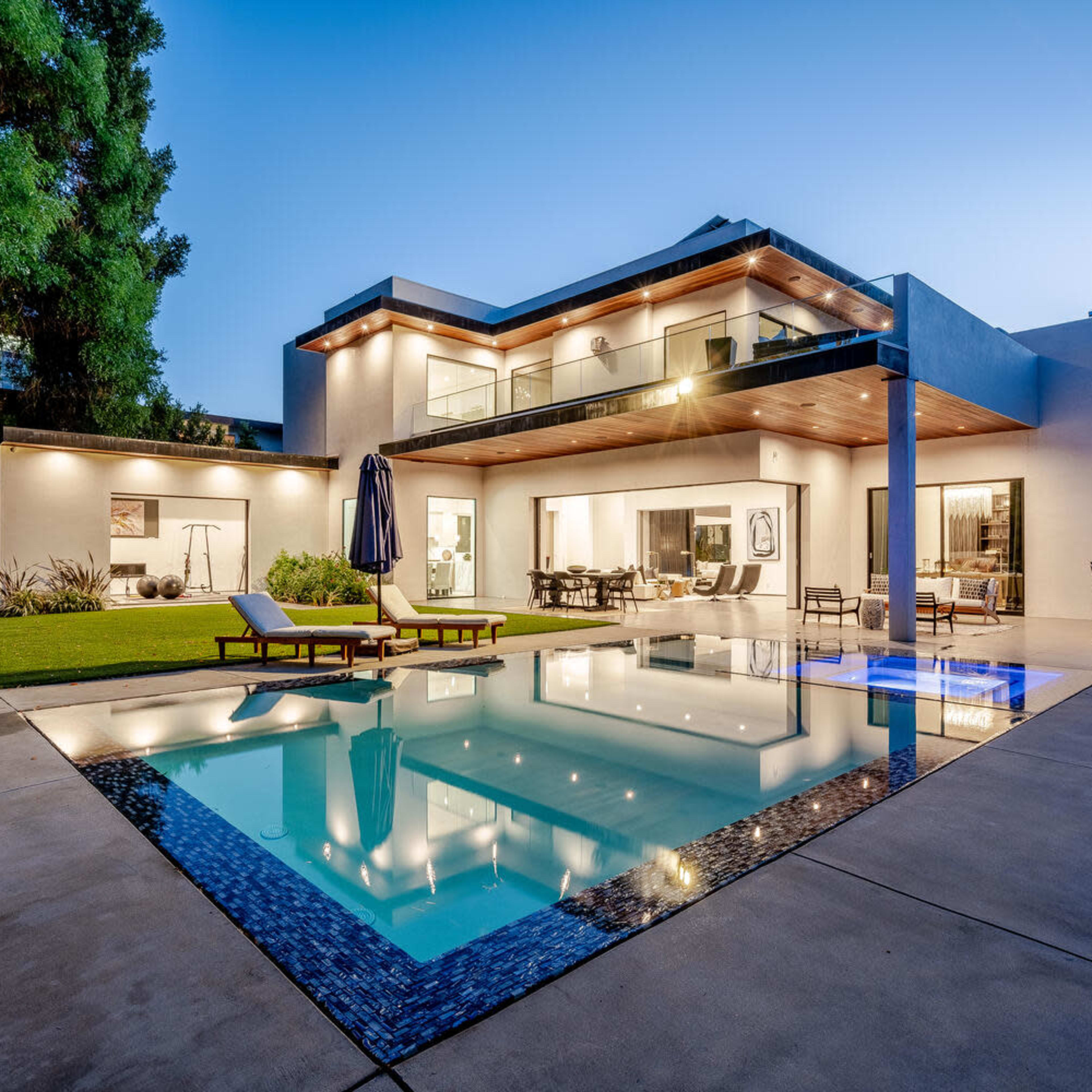 A modern house with large windows and a flat roof surrounds a rectangular pool, featuring lounge chairs and outdoor seating nearby.
