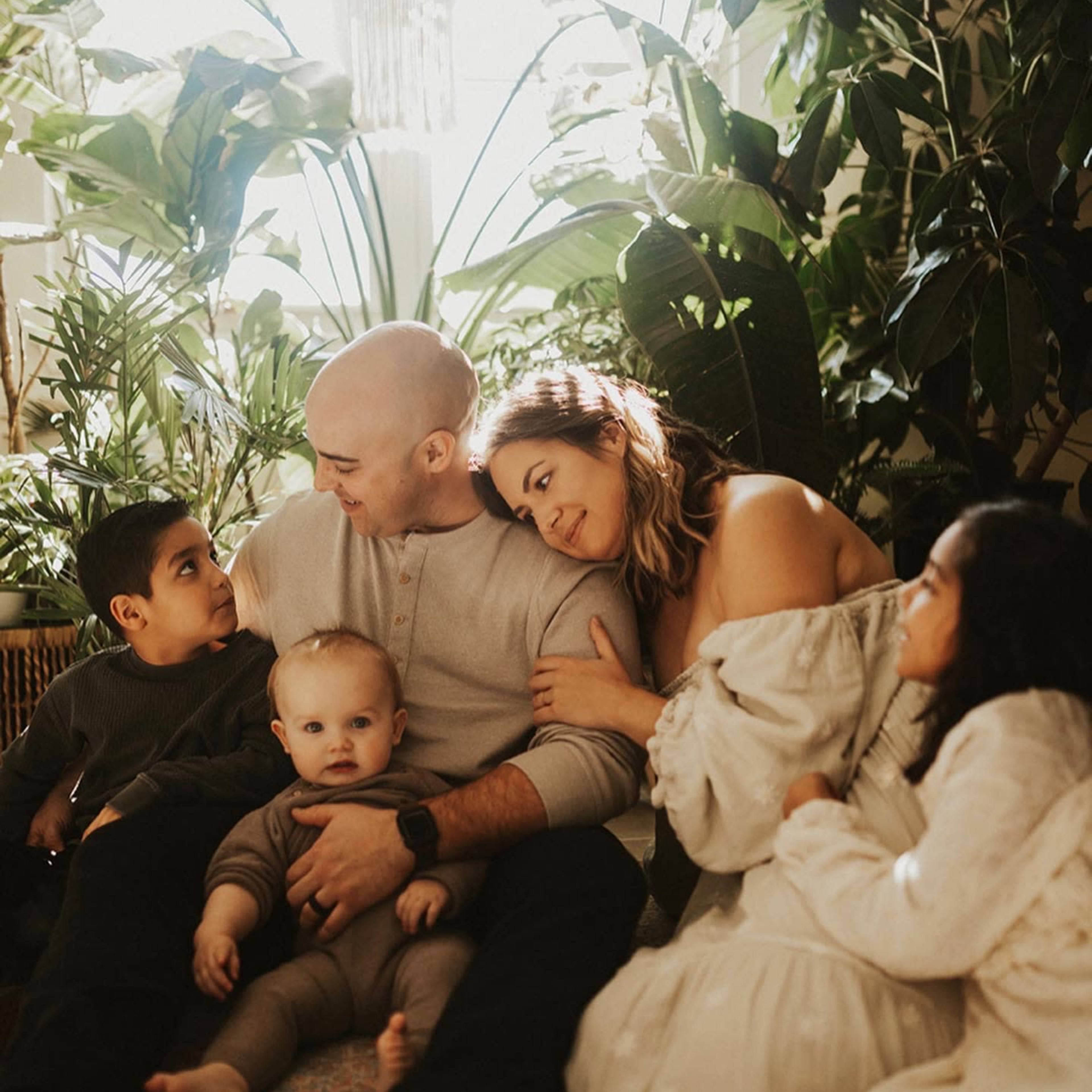A family sits together on the floor surrounded by various indoor plants, with two children looking at their parents and a baby in the mother's lap.