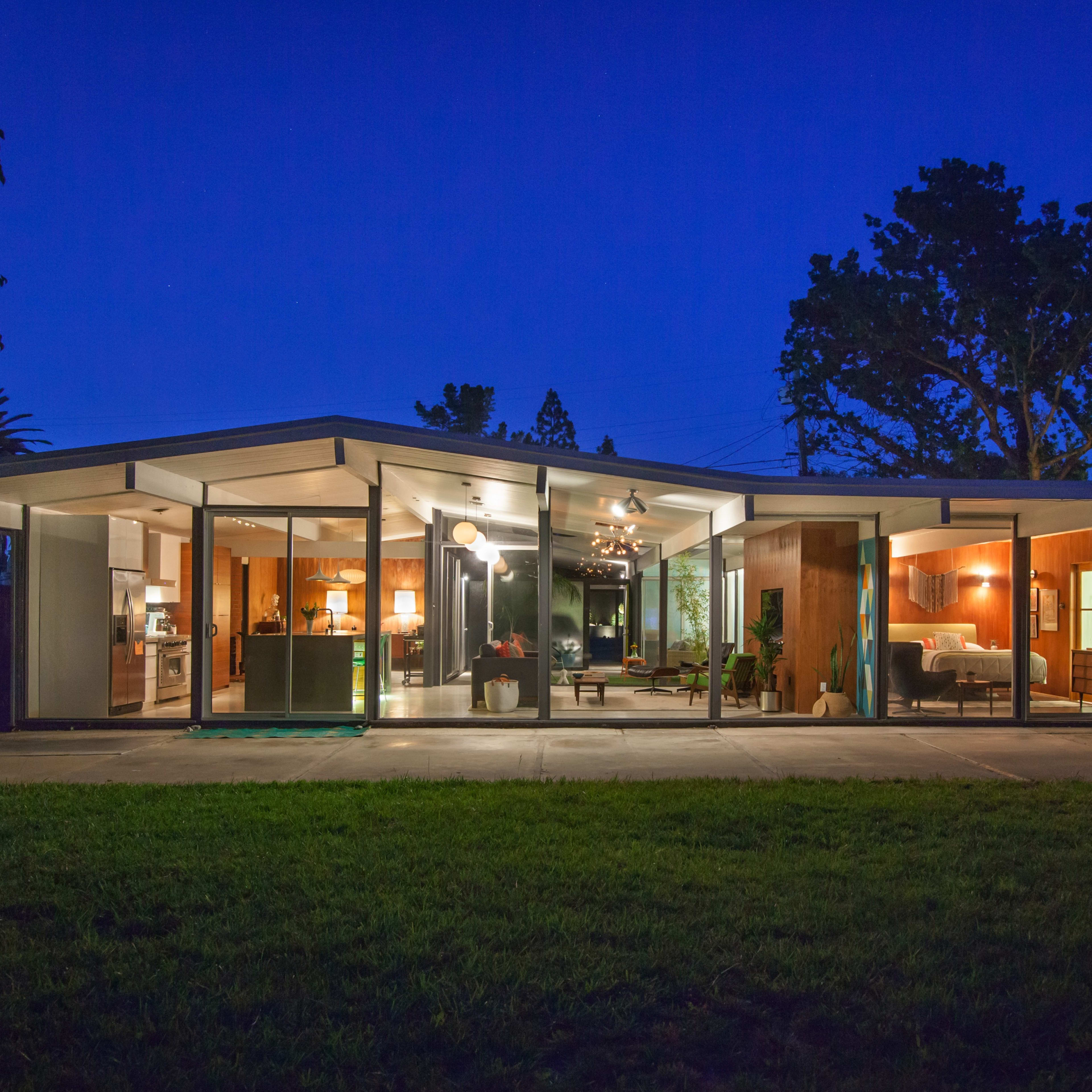 A modern, single-story house with large glass windows is illuminated at night, revealing its interior layout and furnishings.