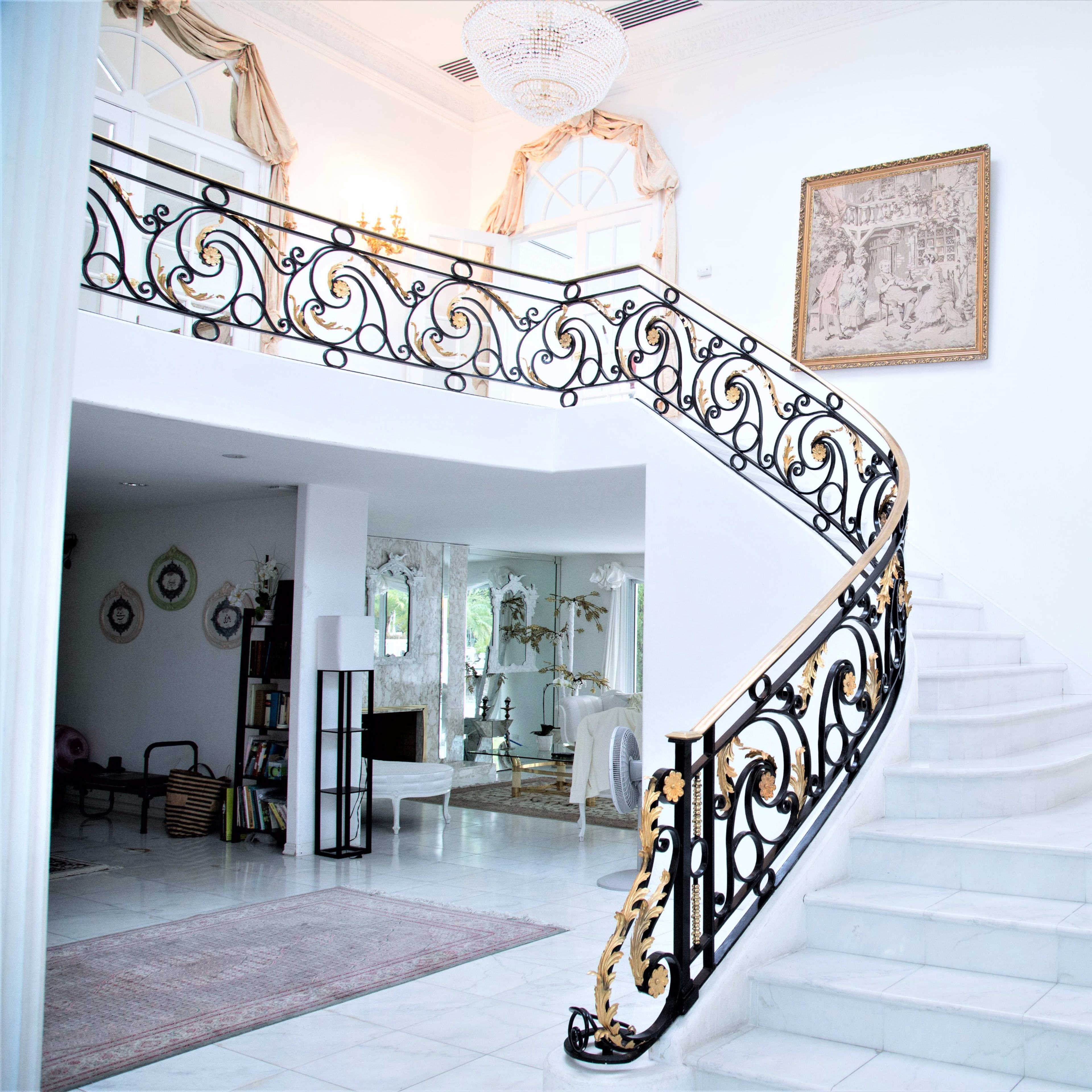 A spacious, elegant foyer with a sweeping staircase featuring ornate black and gold railings, leading to a bright, airy living area.